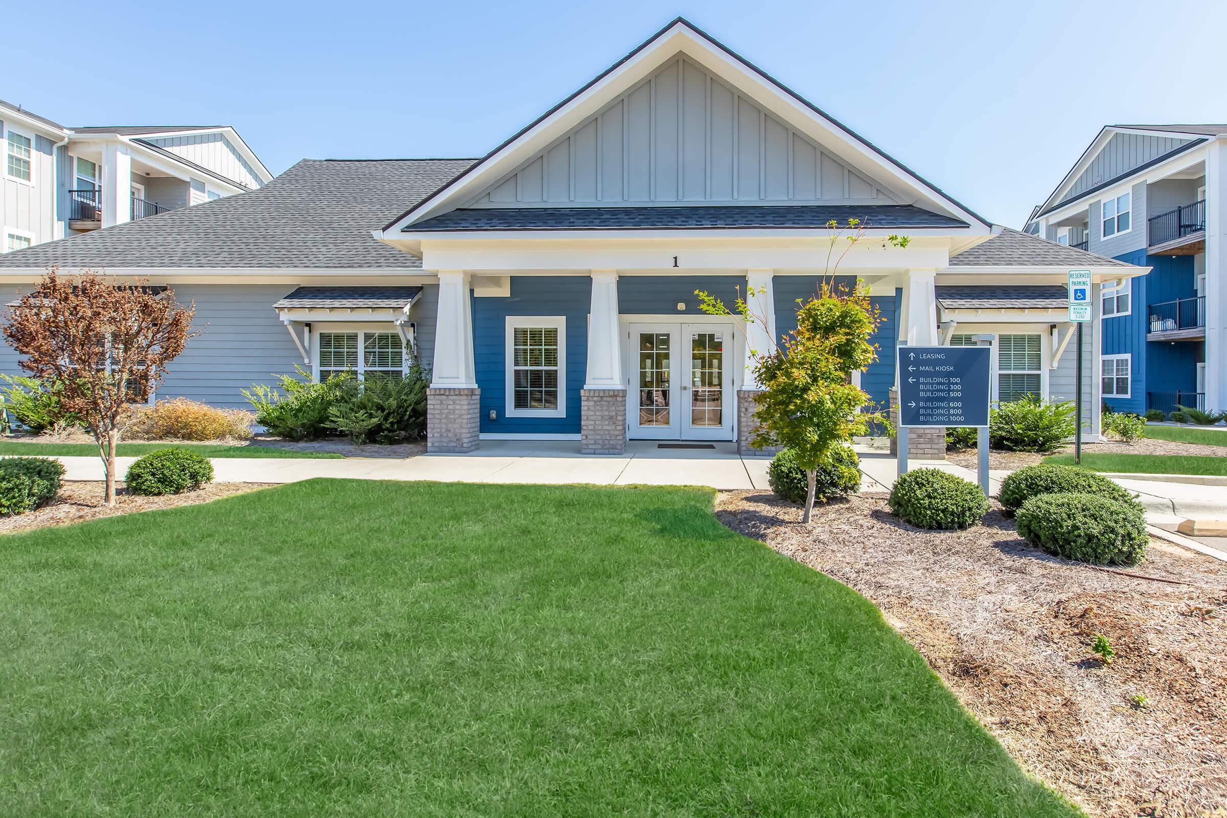 A modern building with a blue and gray exterior, featuring large windows and a welcoming entrance. The surrounding area has well-manicured green grass, neatly trimmed bushes, and a clear blue sky above. Signs near the entrance indicate it is a residential or community facility.