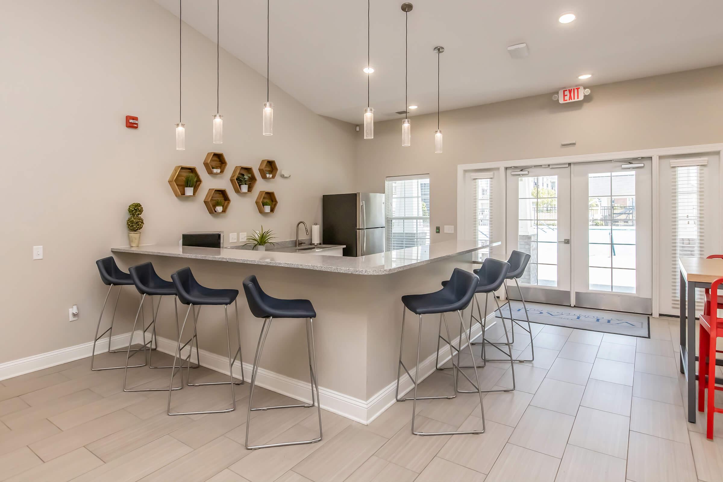 Modern kitchen and dining area with a large island featuring bar seating. Stylish light fixtures hang above. The walls are decorated with hexagonal shelves, and a large window offers a view of the outside. The decor includes potted plants and contemporary appliances, creating a bright and inviting atmosphere.