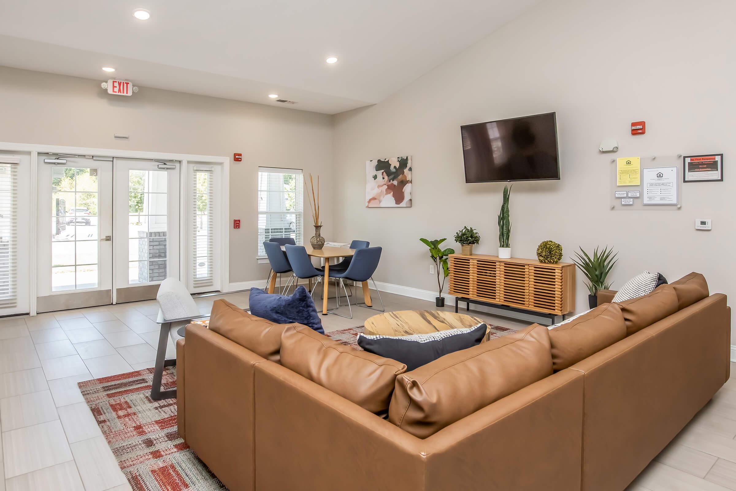 Modern lounge area featuring a large brown leather sectional sofa, a round coffee table, and a dining area with a round table and four blue chairs. A wall-mounted TV is visible, along with decorative plants and artwork. Large windows allow natural light into the space.