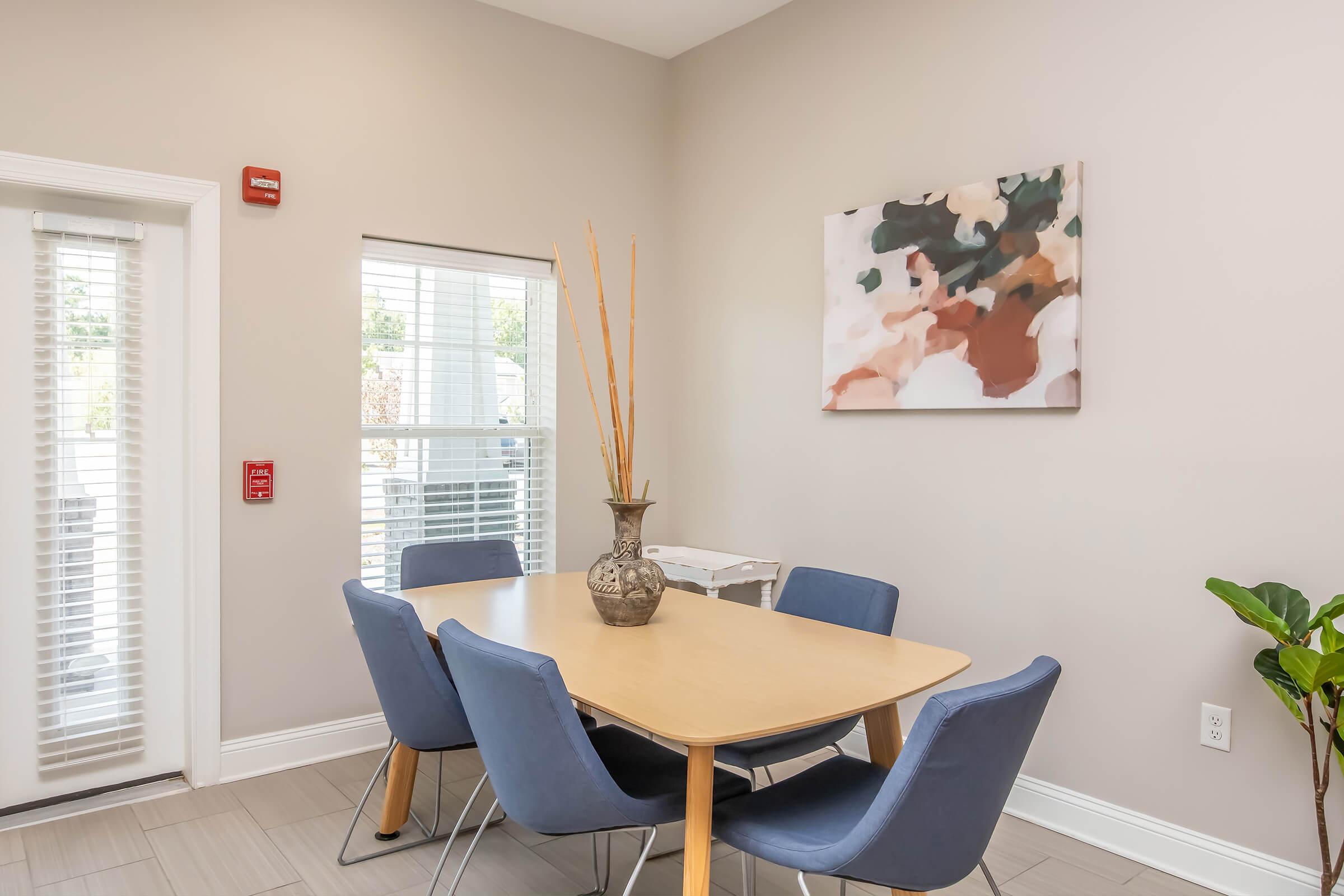 A modern dining area featuring a light wood table surrounded by blue upholstered chairs. A decorative vase with long, slender branches sits at the center of the table. On the wall behind, there is an abstract art piece with soft, muted colors. Natural light streams through the window, adding warmth to the space.