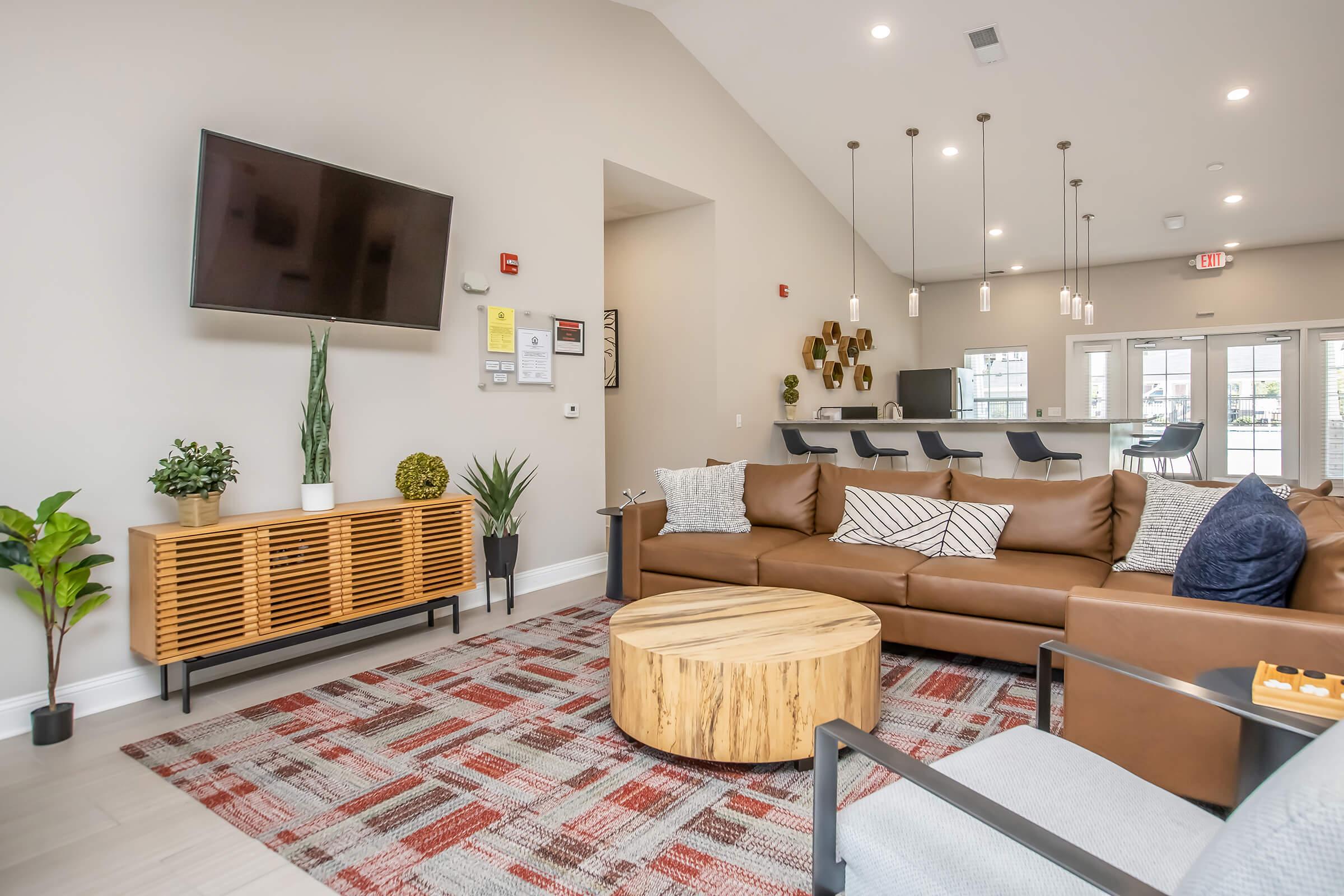 Modern living room featuring a brown leather sofa with decorative pillows, a round wooden coffee table, and a patterned area rug. A flat-screen TV hangs on the wall, and plants add a touch of greenery. The space is well-lit with pendant lighting and has a contemporary design.