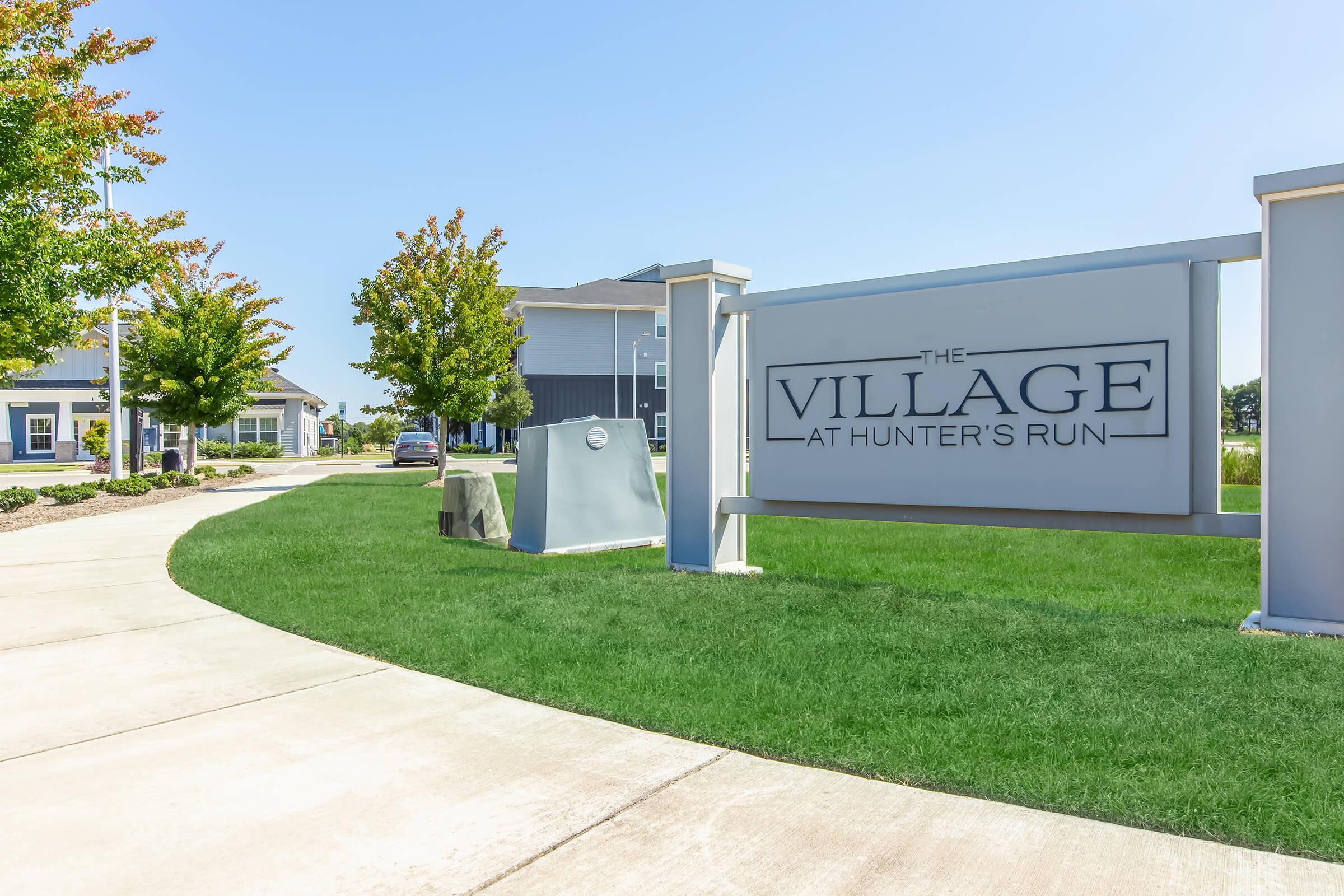 Signage for "The Village at Hunter's Run" with the entrance pathway lined by trees and greenery. A clear blue sky is visible above, and modern buildings can be seen in the background. The scene conveys a welcoming atmosphere for residents and visitors.