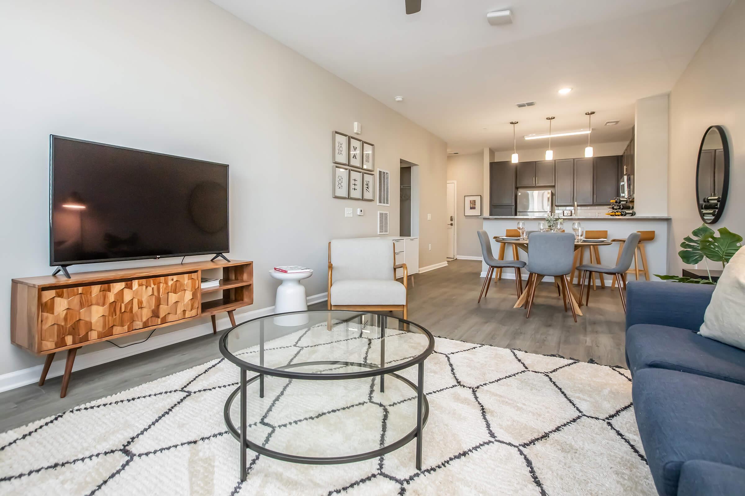Modern living room featuring a flat-screen TV on a wooden cabinet, a round glass coffee table on a patterned rug, and a blue sofa. A dining area with a wooden table and chairs is visible in the background, along with minimalist decor and indoor plants, all in a bright and airy space.