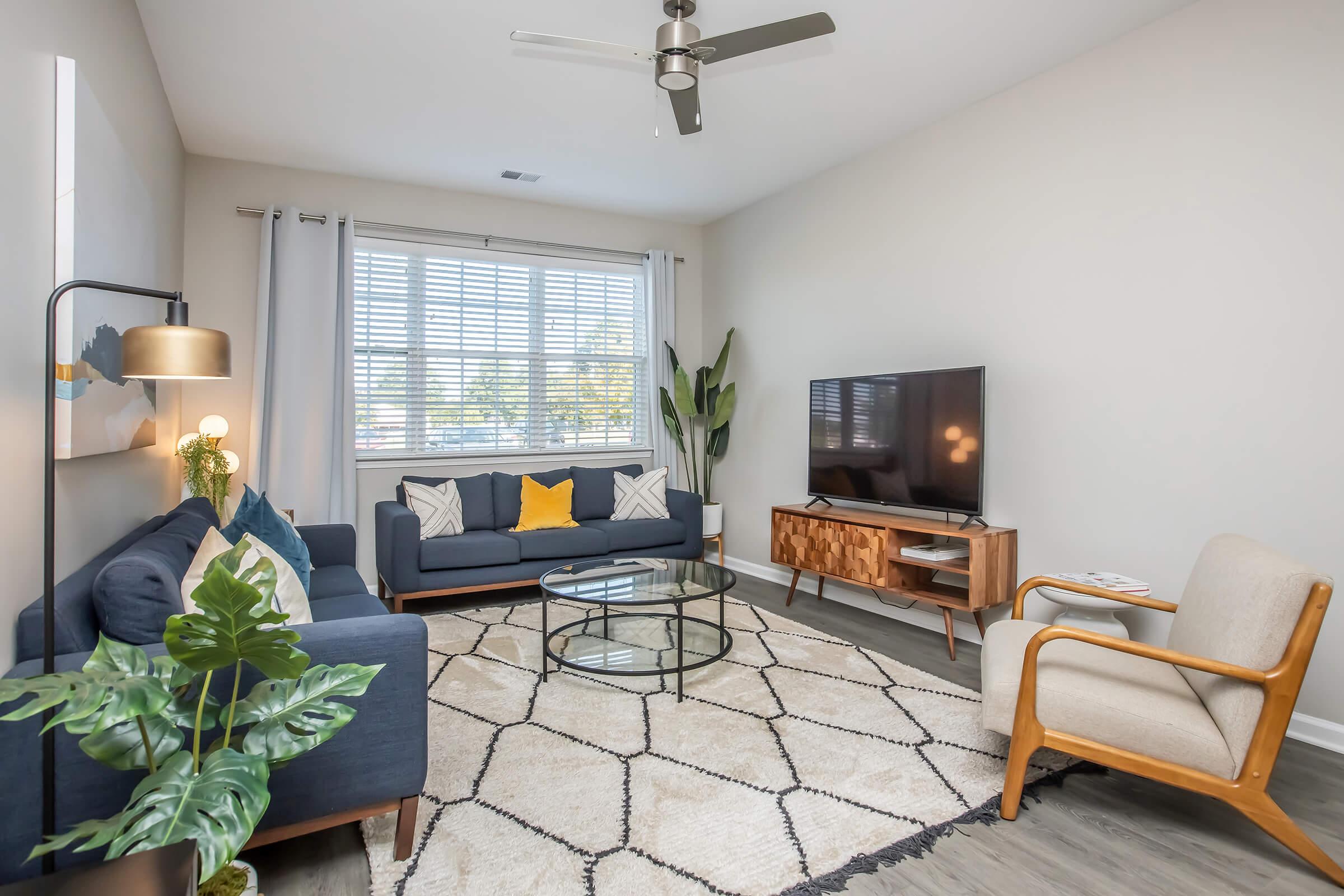 A modern living room featuring two blue sofas with yellow pillows, a round glass coffee table, and a wooden TV stand with a flat-screen TV. A potted plant adds greenery, and the space is illuminated by a stylish floor lamp. Natural light streams in through a large window with white curtains.