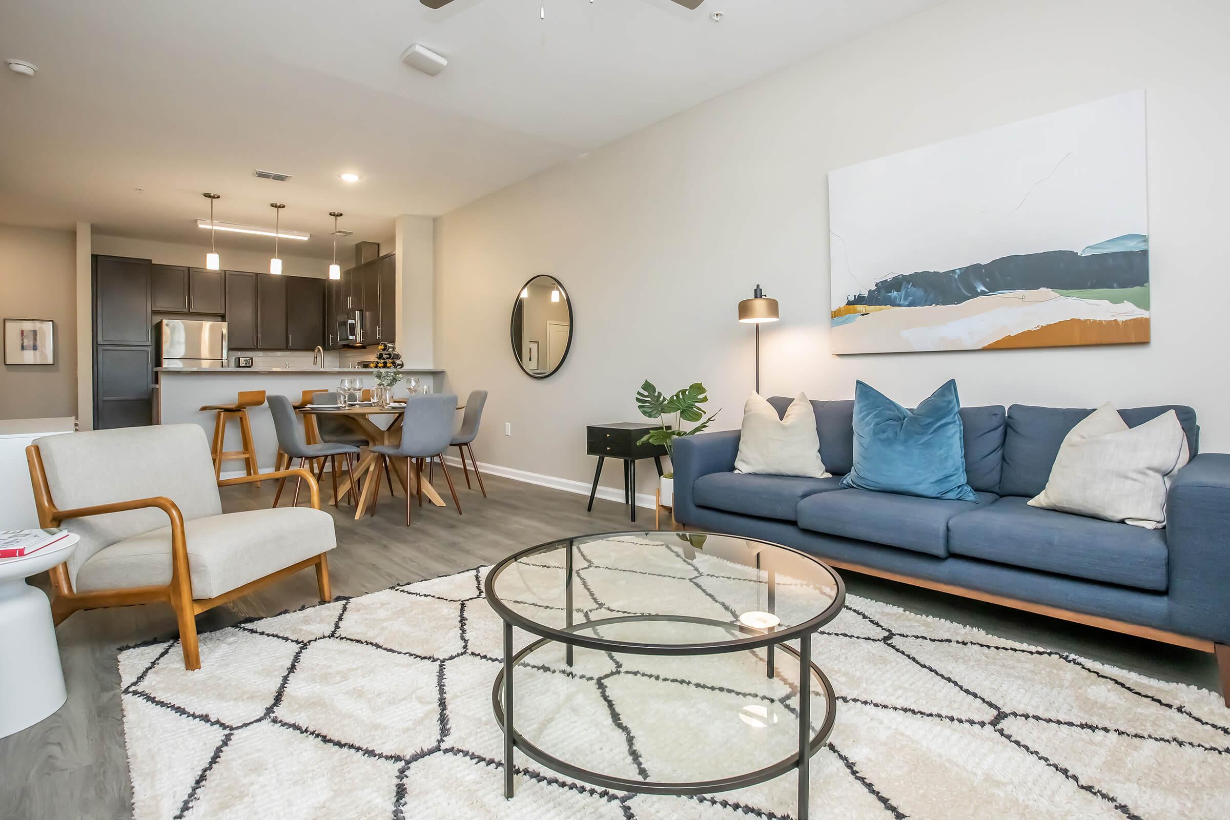 A modern living room featuring a blue couch with decorative pillows, a circular glass coffee table, and a patterned area rug. In the background, there is a dining area with a wooden table and chairs, and a kitchen with dark cabinetry. A large wall art piece adds color to the neutral palette.