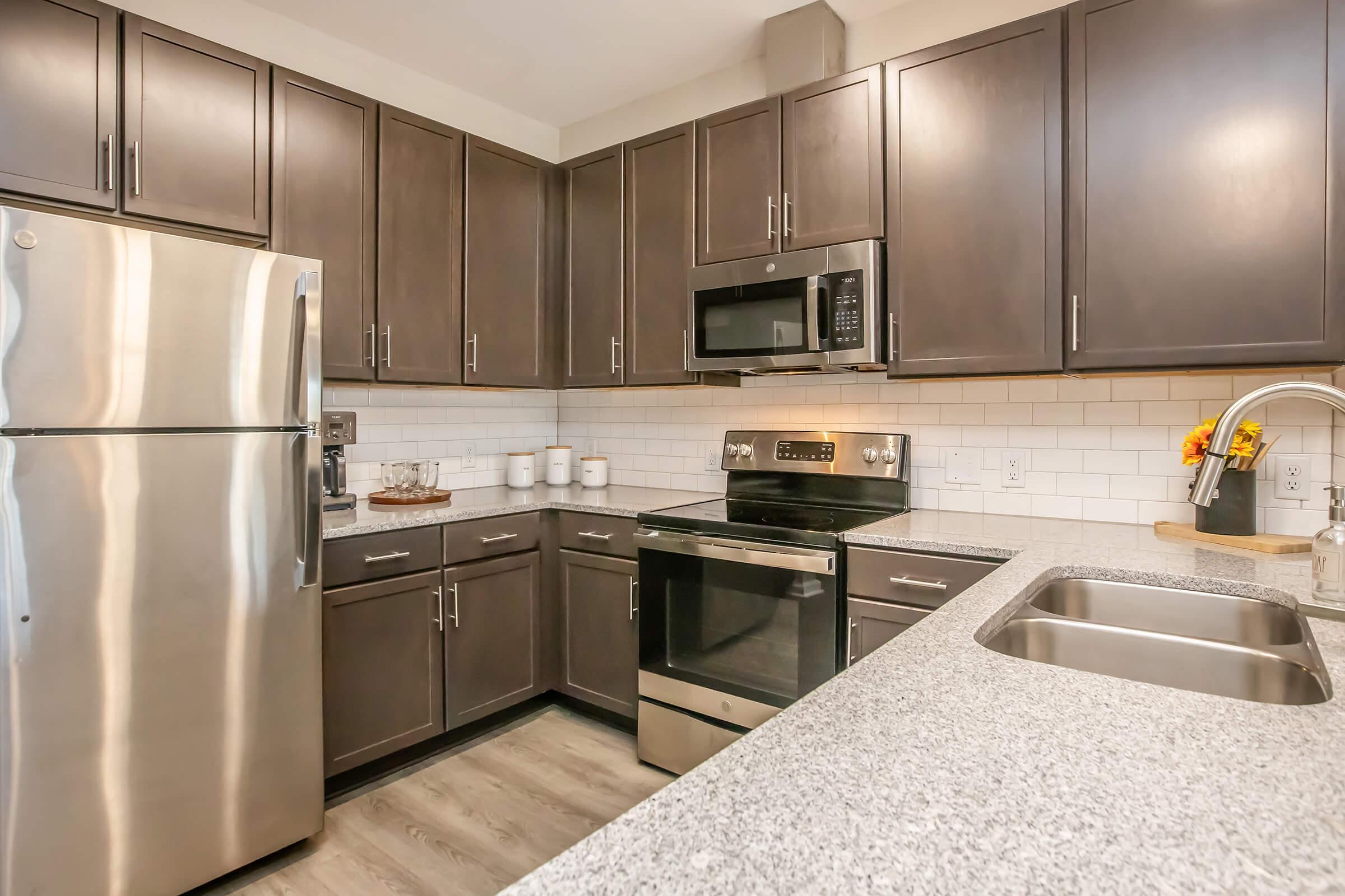 Modern kitchen featuring dark wood cabinetry, stainless steel appliances including a refrigerator, microwave, and oven. The countertop is gray granite with a double sink. White subway tile backsplashes and decorative elements like a flower vase are visible. The space appears clean and well-organized, creating a contemporary atmosphere.