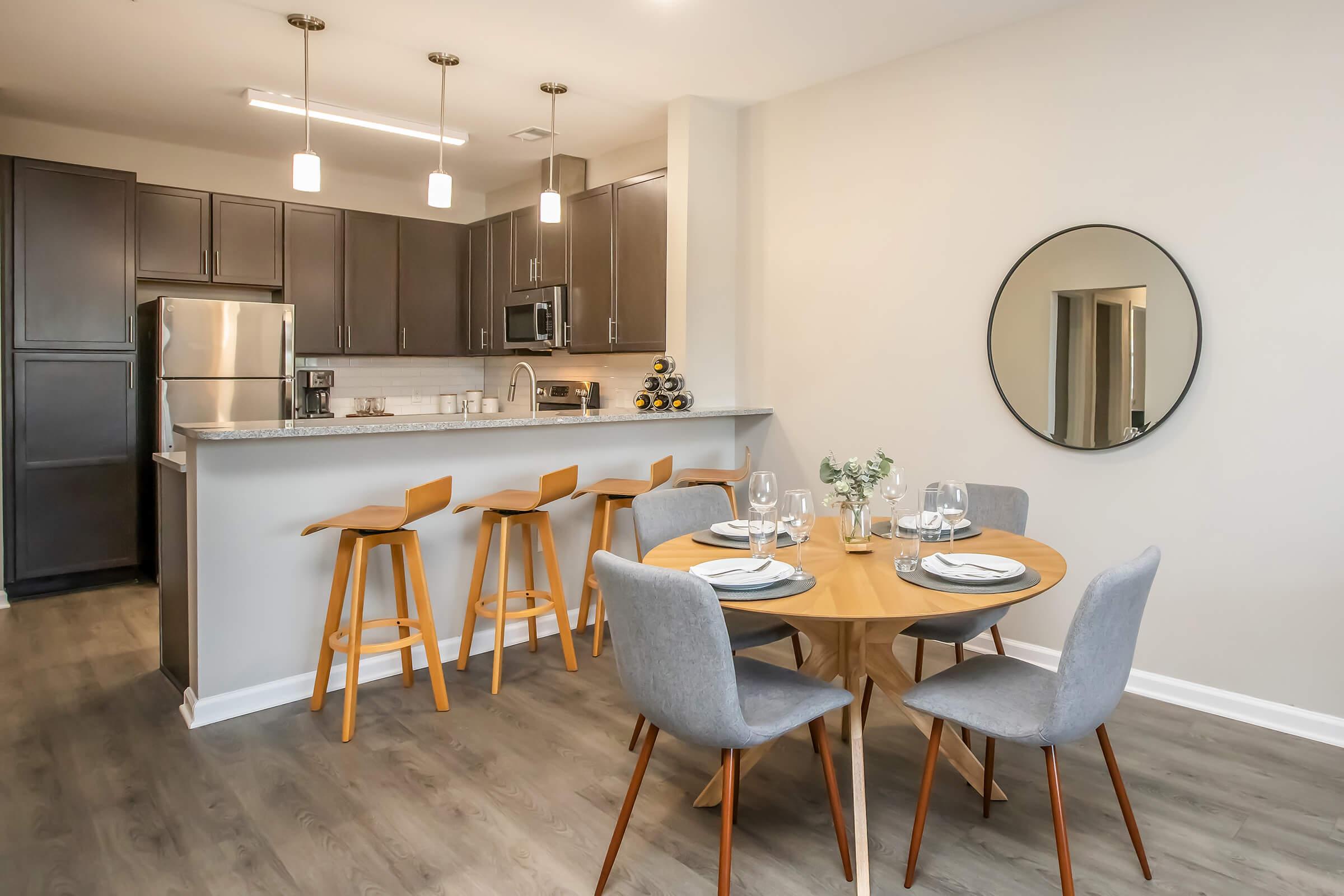 A modern kitchen and dining area featuring a round wooden table with gray chairs, set for four. There are wine glasses and plates on the table. The kitchen has dark cabinets and stainless steel appliances, with pendant lighting above the bar area, and a round mirror on the wall.