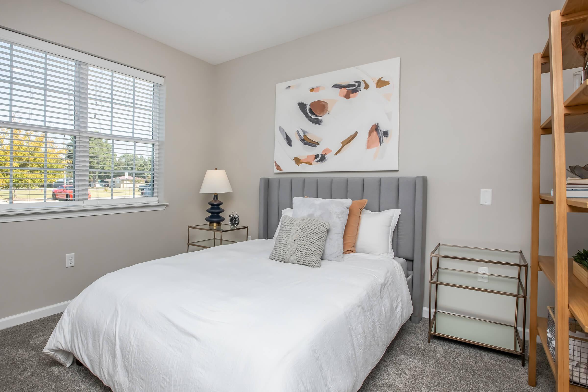 A cozy bedroom featuring a gray upholstered headboard with decorative pillows, a white comforter on the bed, a modern artwork above the bed, and a bedside lamp on a small table. A wooden bookshelf with decorative items is in the corner, and sunlight streams in through a window with white blinds.