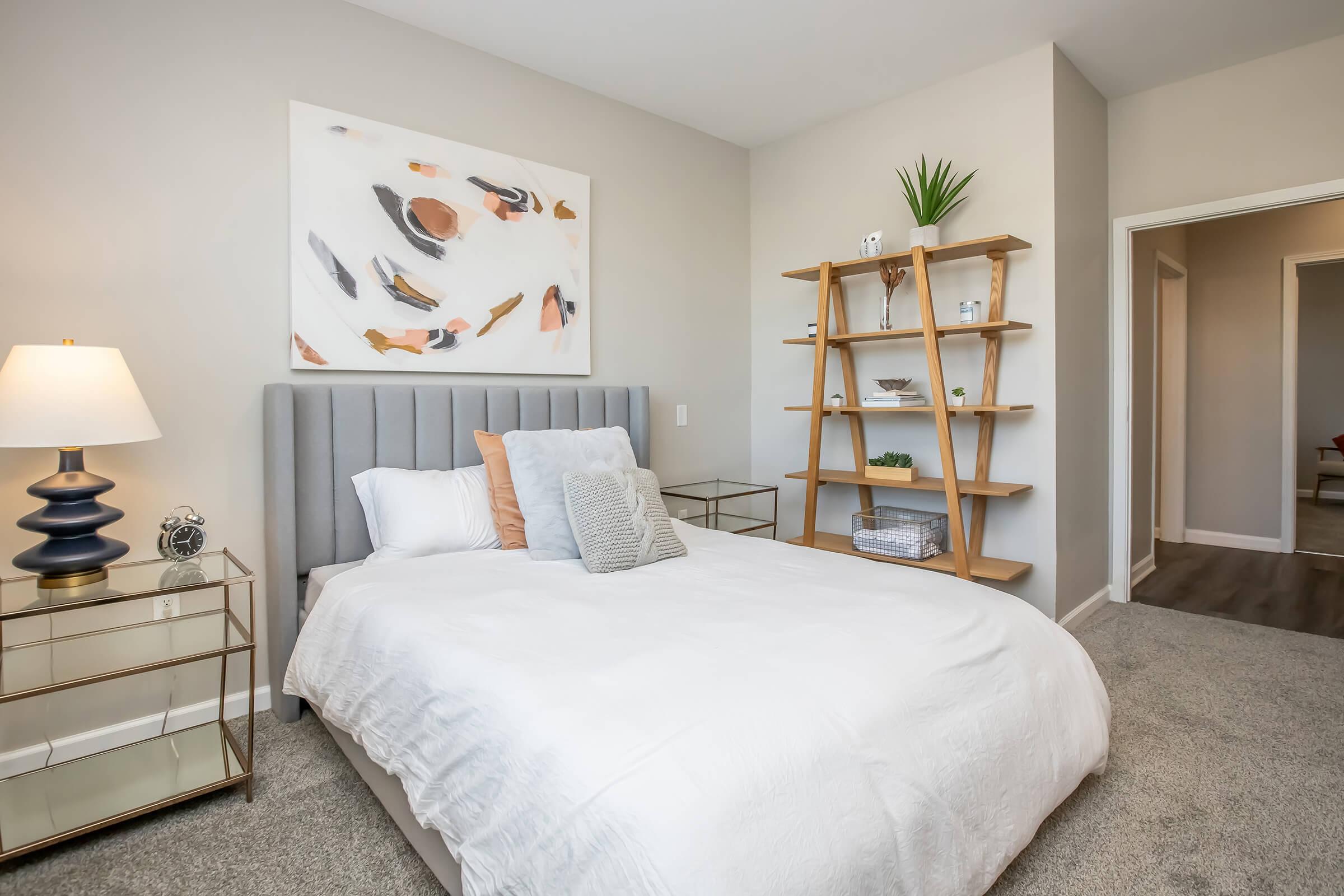 A cozy bedroom featuring a gray upholstered bed with a white bedspread, decorative pillows, and a minimalist decorative shelf. The room has a stylish lamp on a nightstand, an abstract painting on the wall, and soft carpeted flooring. A doorway leads to another room.