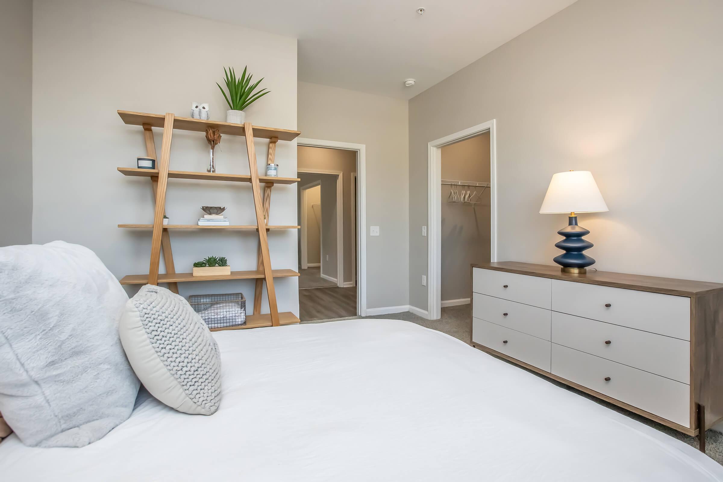 Bright and modern bedroom featuring a neatly made bed with decorative pillows, a wooden shelving unit with plants and decorative items, and a stylish dresser. Soft neutral wall colors enhance the spacious feel, while a lamp provides warm lighting. Two open doorways lead to additional rooms.