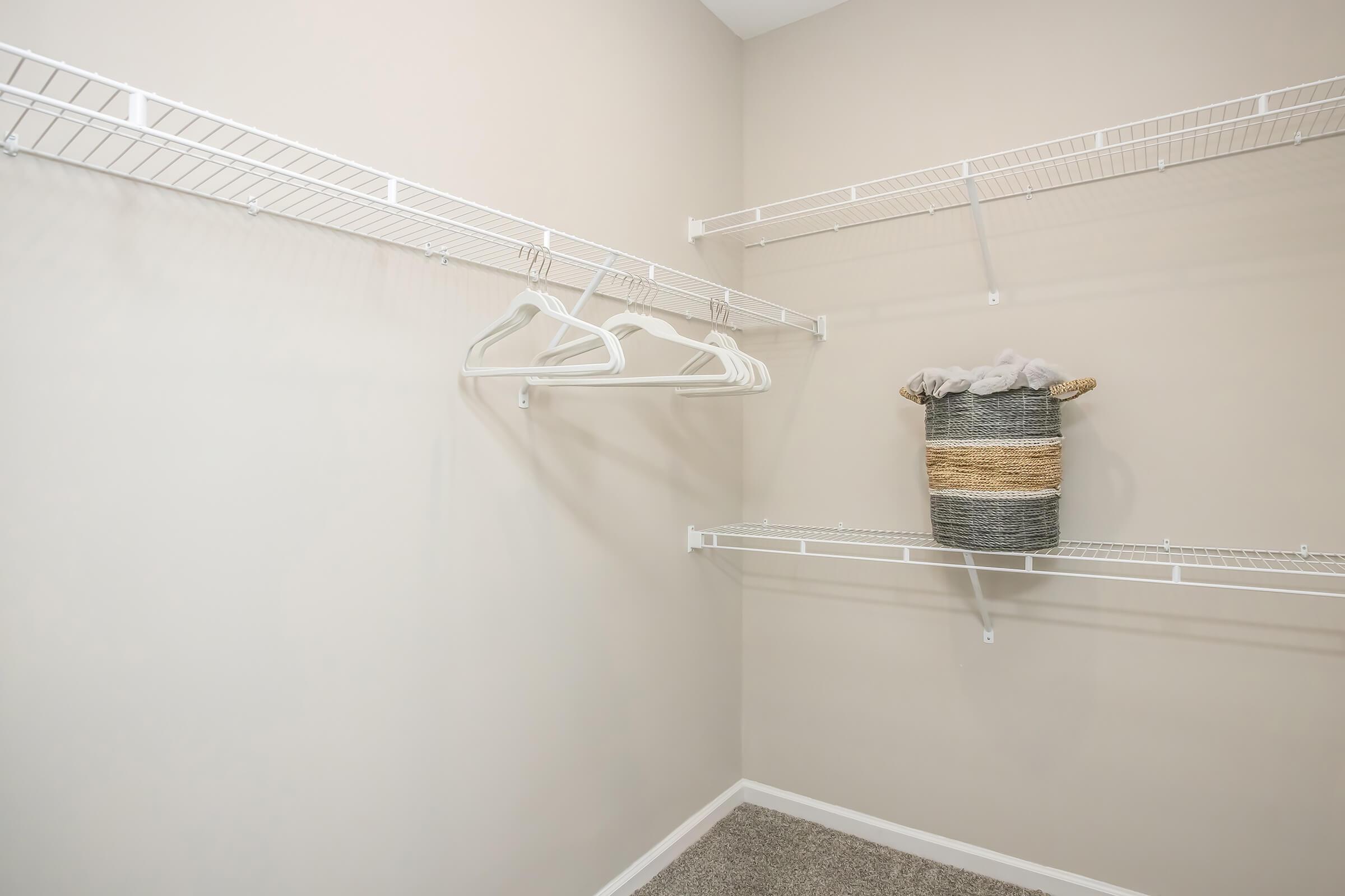A clean and organized closet featuring white wire shelving. There are several empty white hangers hanging from the shelves, and a woven basket sits on the middle shelf, suggesting an uncluttered and tidy storage space. The walls are painted a neutral beige, creating a calm atmosphere.