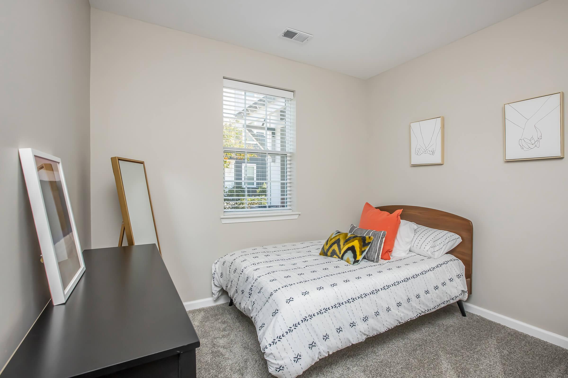 Cozy bedroom featuring a bed with patterned bedding and decorative pillows, a mirror, and a simple dresser. The room has light-colored walls, a window with blinds letting in natural light, and minimalist framed artwork on the walls, creating a warm and inviting atmosphere.