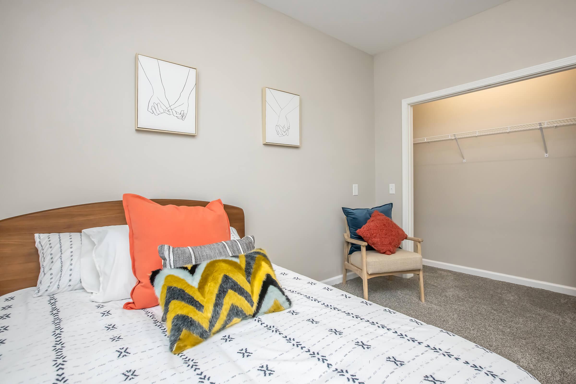 A cozy bedroom featuring a double bed with patterned bedding and colorful throw pillows. Two minimalist artworks of hands in frames hang on the wall above the bed. An empty closet with a shelf is visible on the right, and a comfortable chair with a blue and red pillow sits nearby on a carpeted floor.