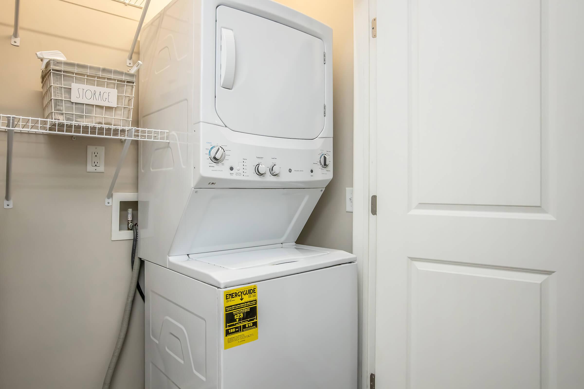 A compact stacked washer and dryer unit in a small laundry space with beige walls. There is a white shelf labeled "STORAGE" above the appliances, and a door on the right side. The dryer has a warning label on it, and the area appears organized and well-lit.