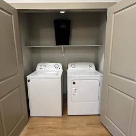 A small laundry closet featuring a white washing machine and a dryer side by side. Above them is a shelf, and a black laundry basket is placed on the shelf. The closet has beige walls and double doors that are partially open, revealing the appliances and storage space inside.