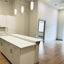 A modern, vacant room featuring a white kitchen countertop with cabinets, pendant lighting, and a spacious area with wooden flooring. There is a large window allowing natural light, and a doorway leading to another room. The walls are light-colored, creating an open and airy atmosphere.