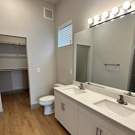 Spacious modern bathroom featuring a double vanity with two sinks, a large mirror, and stylish lighting. The room includes a toilet and a closet with ample storage space. The flooring is wood-like, and there is a window allowing natural light to enter. The color scheme is neutral and contemporary.