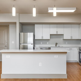 Modern kitchen featuring an open layout with white cabinetry, stainless steel appliances, a large island with a wooden countertop, and sleek pendant lighting. The space has light hardwood flooring and neutral wall colors, creating a bright and inviting atmosphere.