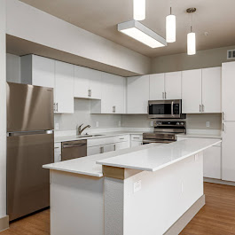 Modern kitchen featuring white cabinets, stainless steel refrigerator, and microwave. The island has a white countertop with a built-in sink. Bright lighting from pendant fixtures adds to the spacious, contemporary design with hardwood flooring.