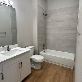 A modern bathroom featuring a white bathtub with a shower, a sleek vanity with a sink, a large mirror above the vanity, and a toilet. The walls are adorned with light gray tiles, and the floor has a warm wood finish. Natural light enhances the clean, minimalist design.