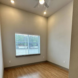 A well-lit, empty room with light gray walls and a wooden floor. It features a large window with blinds and a ceiling fan. The space has a modern, minimalist design, making it versatile for various uses.