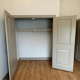 A spacious, empty closet with two open gray doors, featuring a single clothing rod and light-colored walls. The floor is made of light wood, providing a clean and modern look.