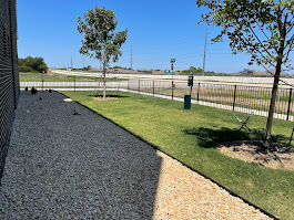 A landscaped outdoor area featuring a patch of green grass bordered by gravel. In the background, there is a fenced area and an open road. A few small trees are present, and the sky is clear and blue.