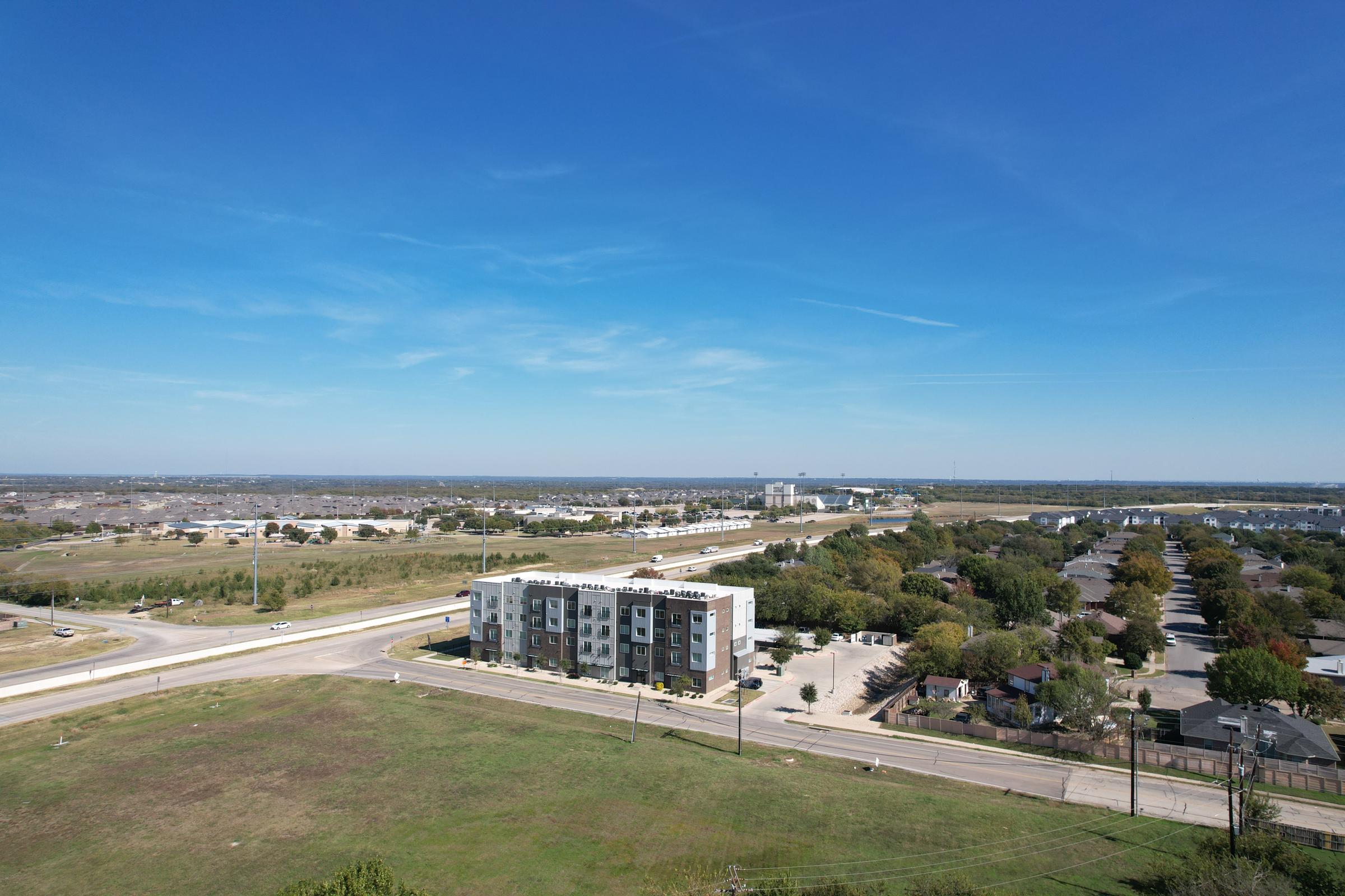 Aerial view of a suburban landscape featuring a multi-story apartment building on the left, flanked by a road and open grassy area. In the background, small residential homes are visible along the road, with a clear blue sky above and distant structures on the horizon.