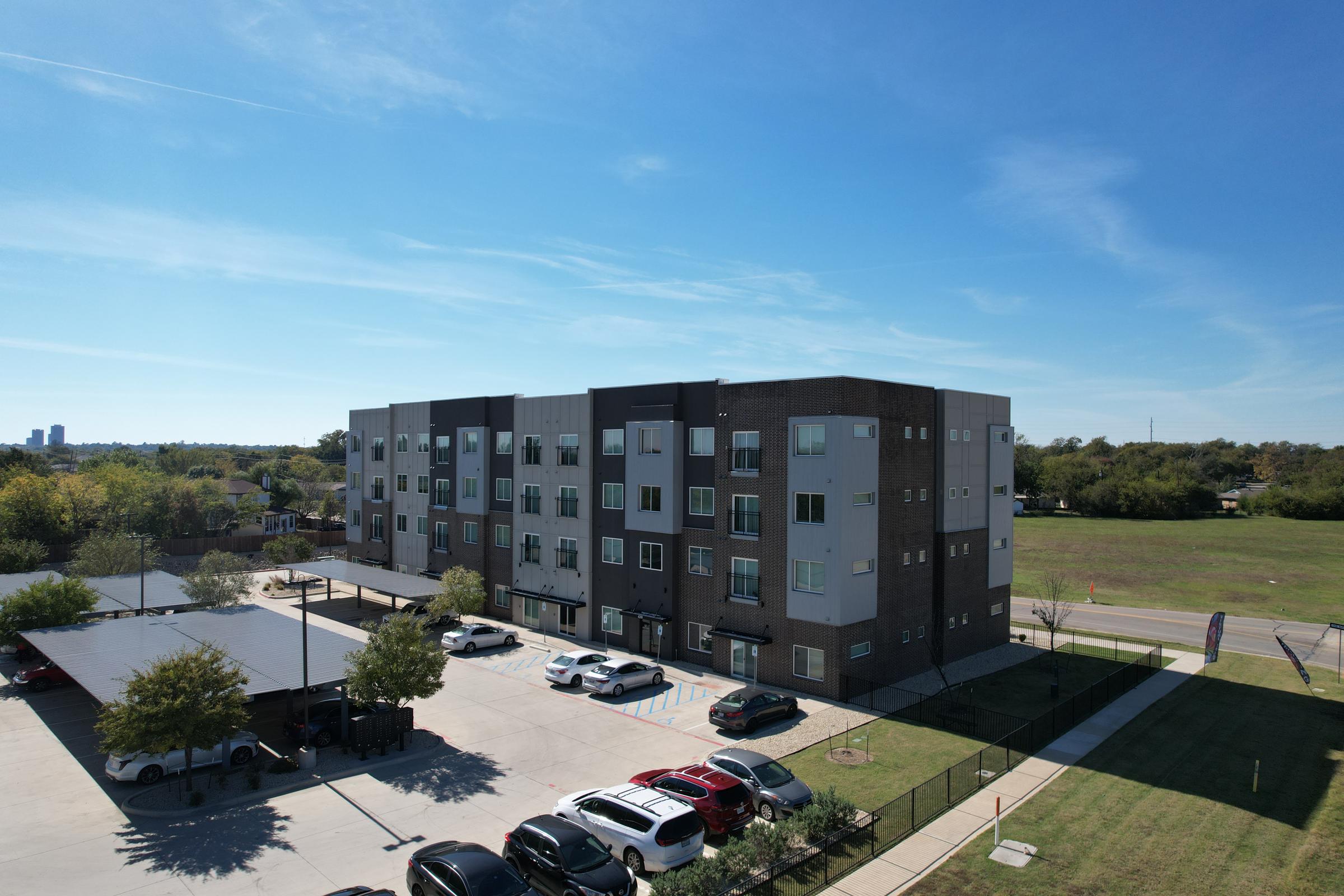 Modern apartment building with multiple floors, featuring a mix of large windows and balcony spaces. In the foreground, there is a parking area with several parked cars. The background showcases green grass and trees, under a clear blue sky.