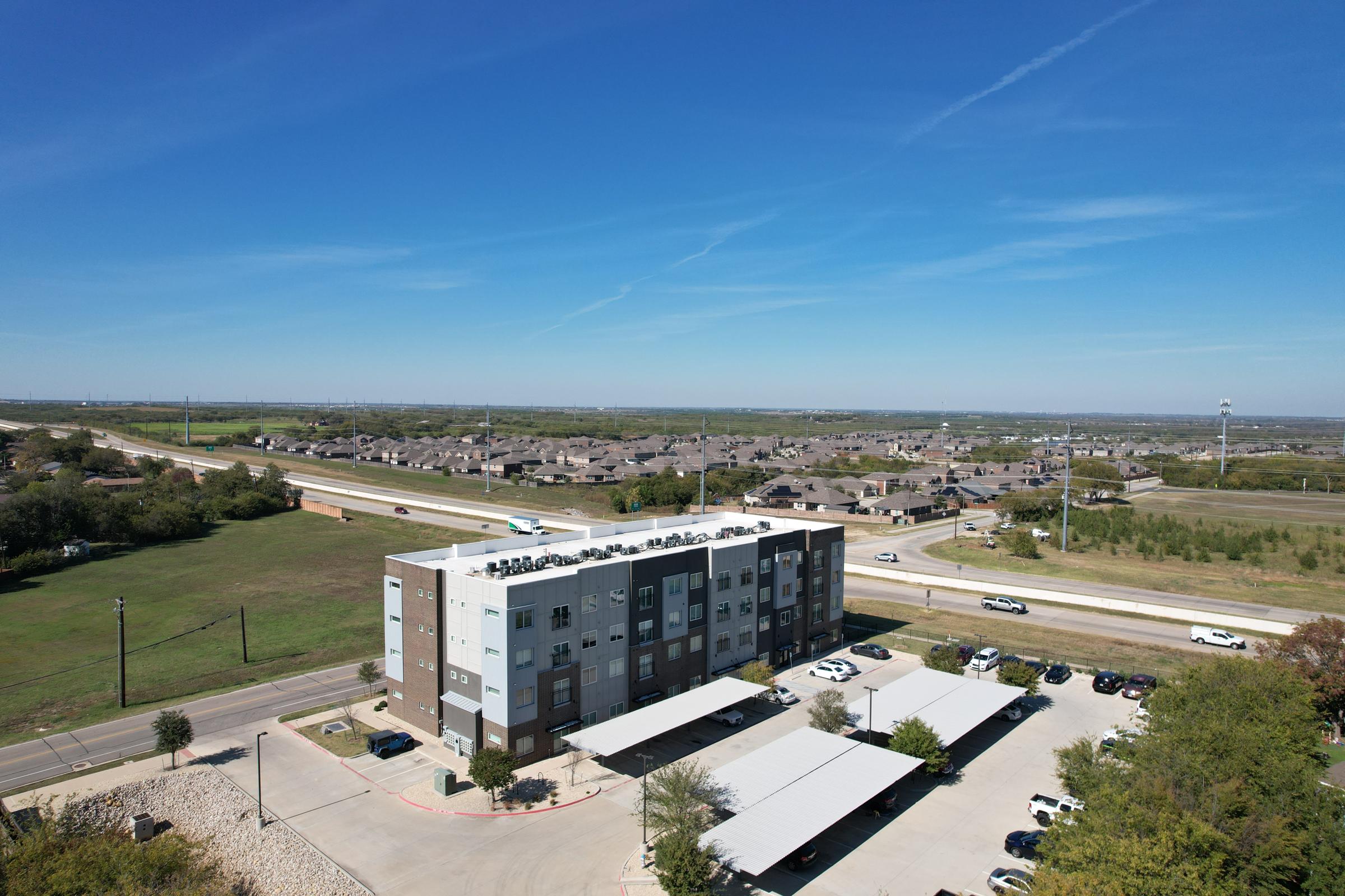 Aerial view of a modern multi-story building with solar panels on the roof, surrounded by a parking lot and covered carports. In the background, a highway runs alongside residential homes and green fields under a clear blue sky.