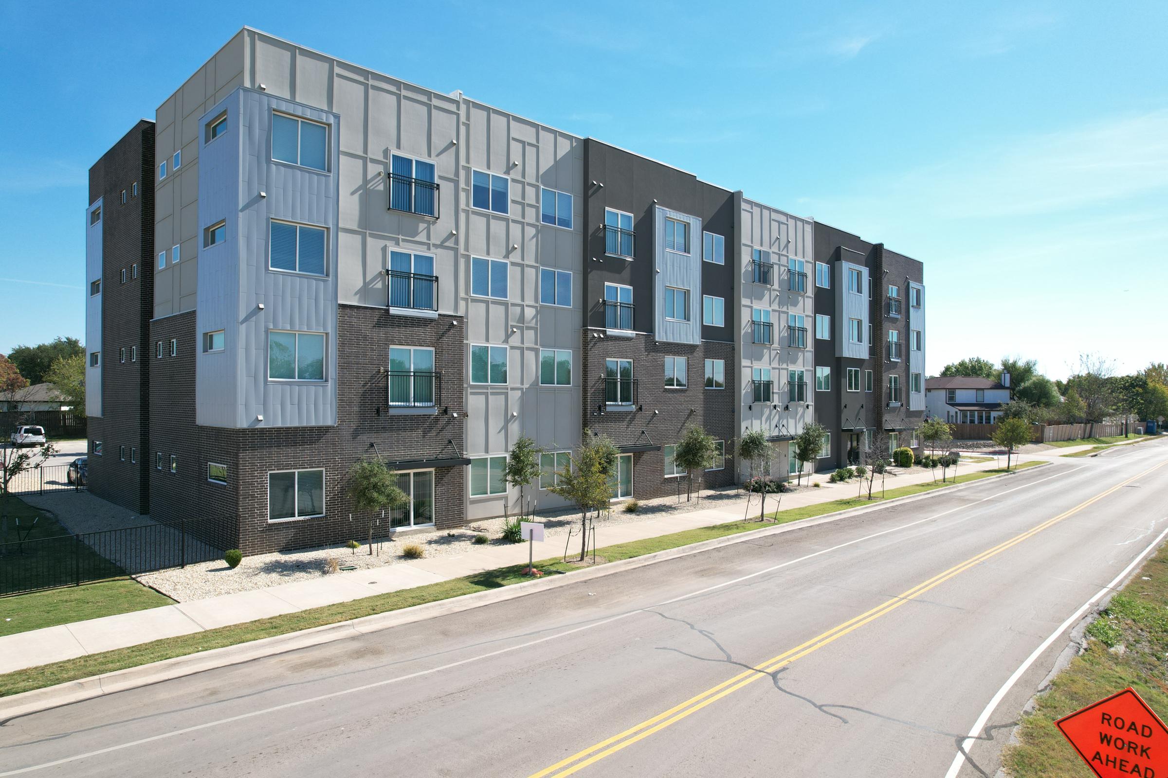 Modern residential apartment building with multiple stories, featuring large windows and a mix of gray and brown exterior. The structure is lined with small trees and shrubbery along a sidewalk, with a road in front marked by a "Road Work Ahead" sign. A clear blue sky is visible overhead.