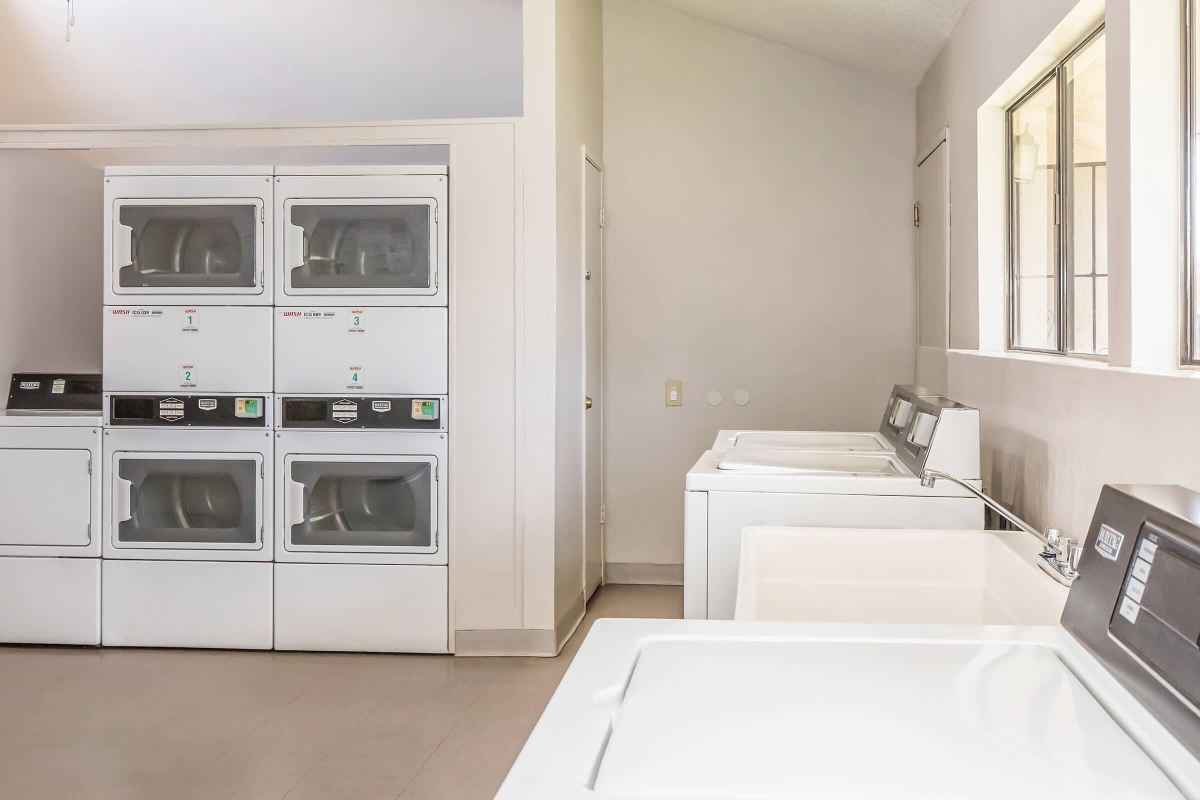 A bright laundry room featuring several washing machines and dryers. There are two stacked dryer units on the left and a row of washing machines on the right. Large windows allow natural light to fill the space, highlighting the clean and organized setting. The walls are painted in a soft, neutral color.