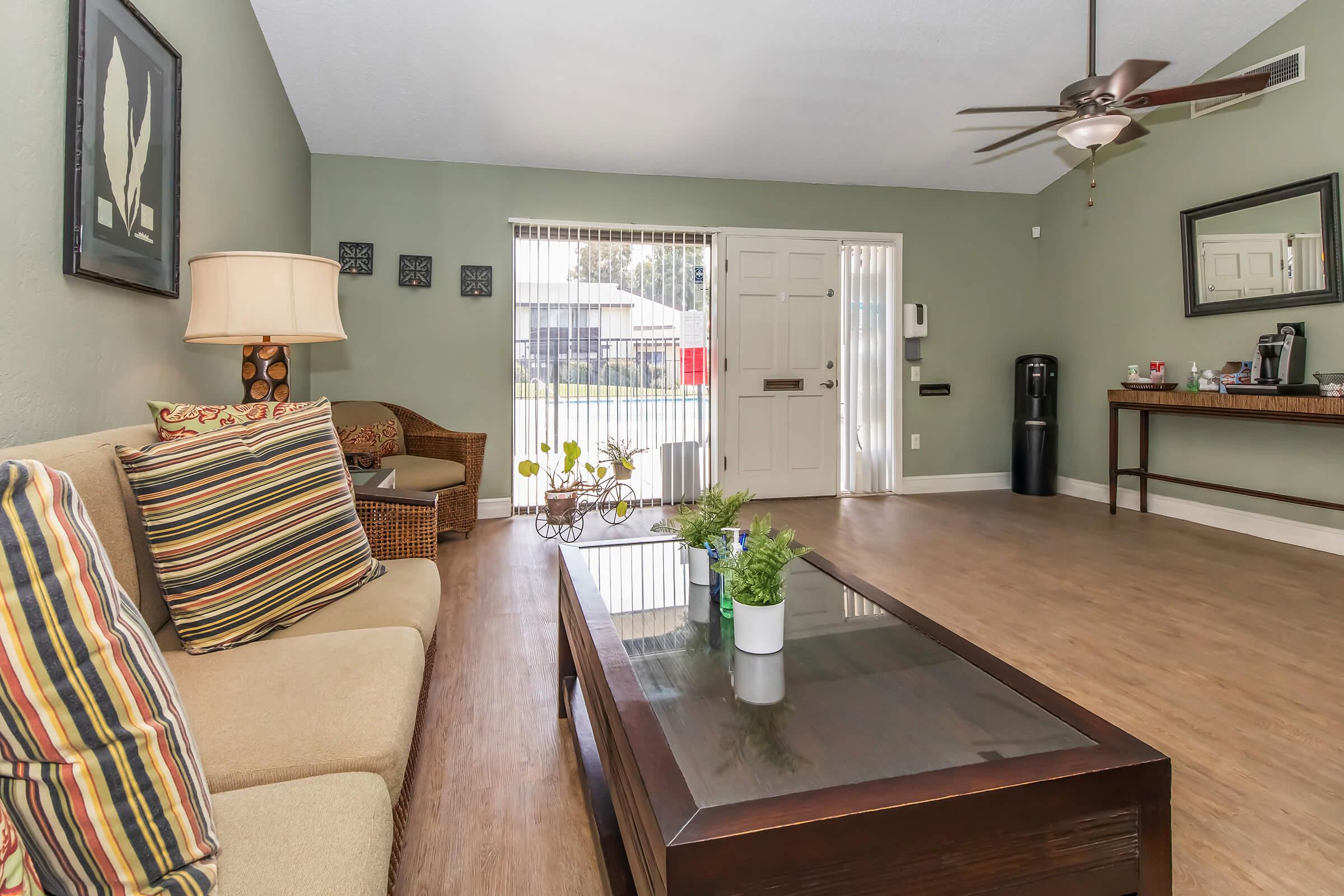 A cozy living room featuring a beige sectional sofa with striped cushions, a wooden coffee table with small potted plants, and a decorative lamp. The space has green walls, a ceiling fan, and large sliding doors leading to an outdoor area. A beverage station and a black water dispenser are visible.