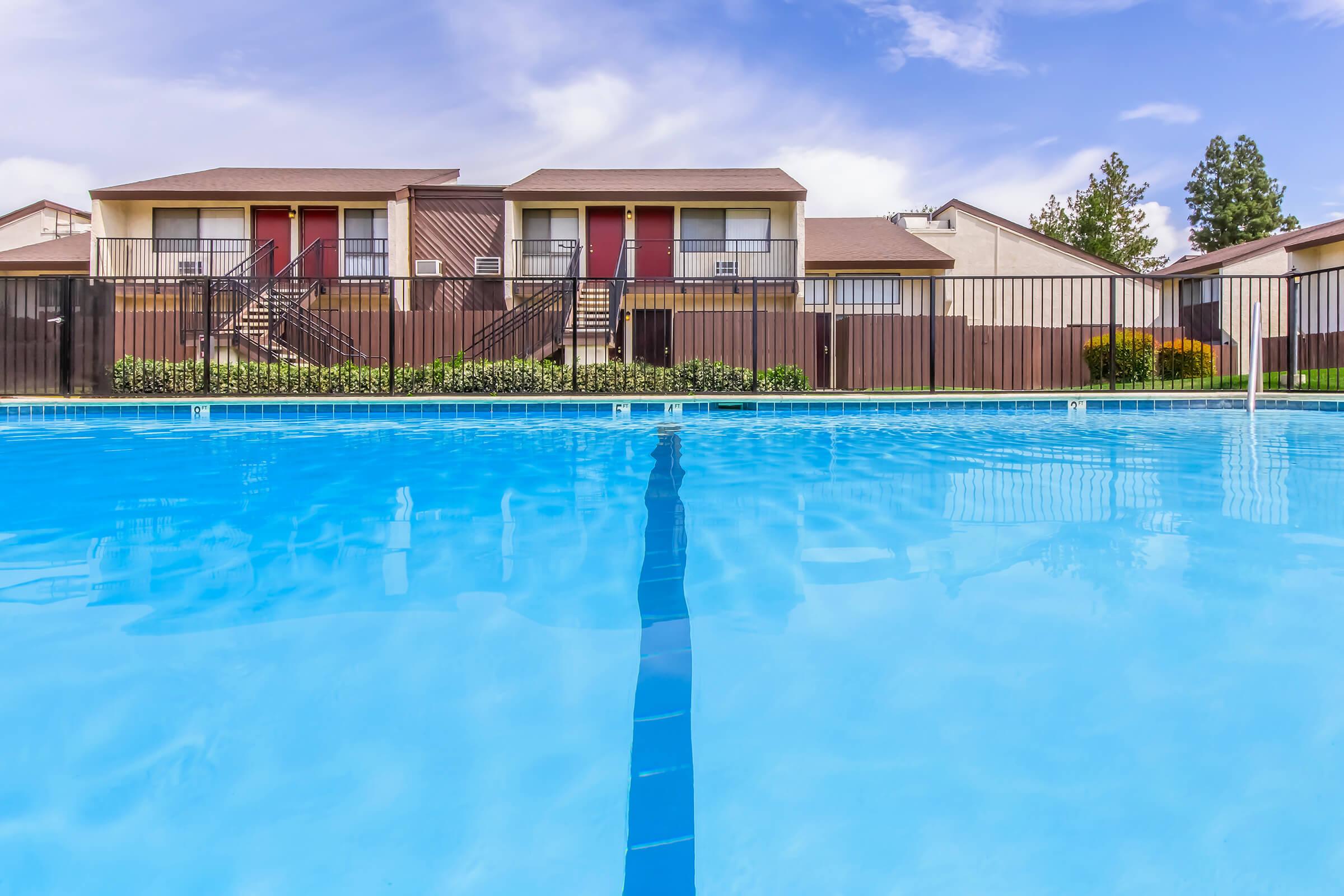 Clear blue swimming pool with a reflection of a two-story apartment building in the background. The surrounding area features a fenced enclosure, green grass, and a clear blue sky with some clouds.