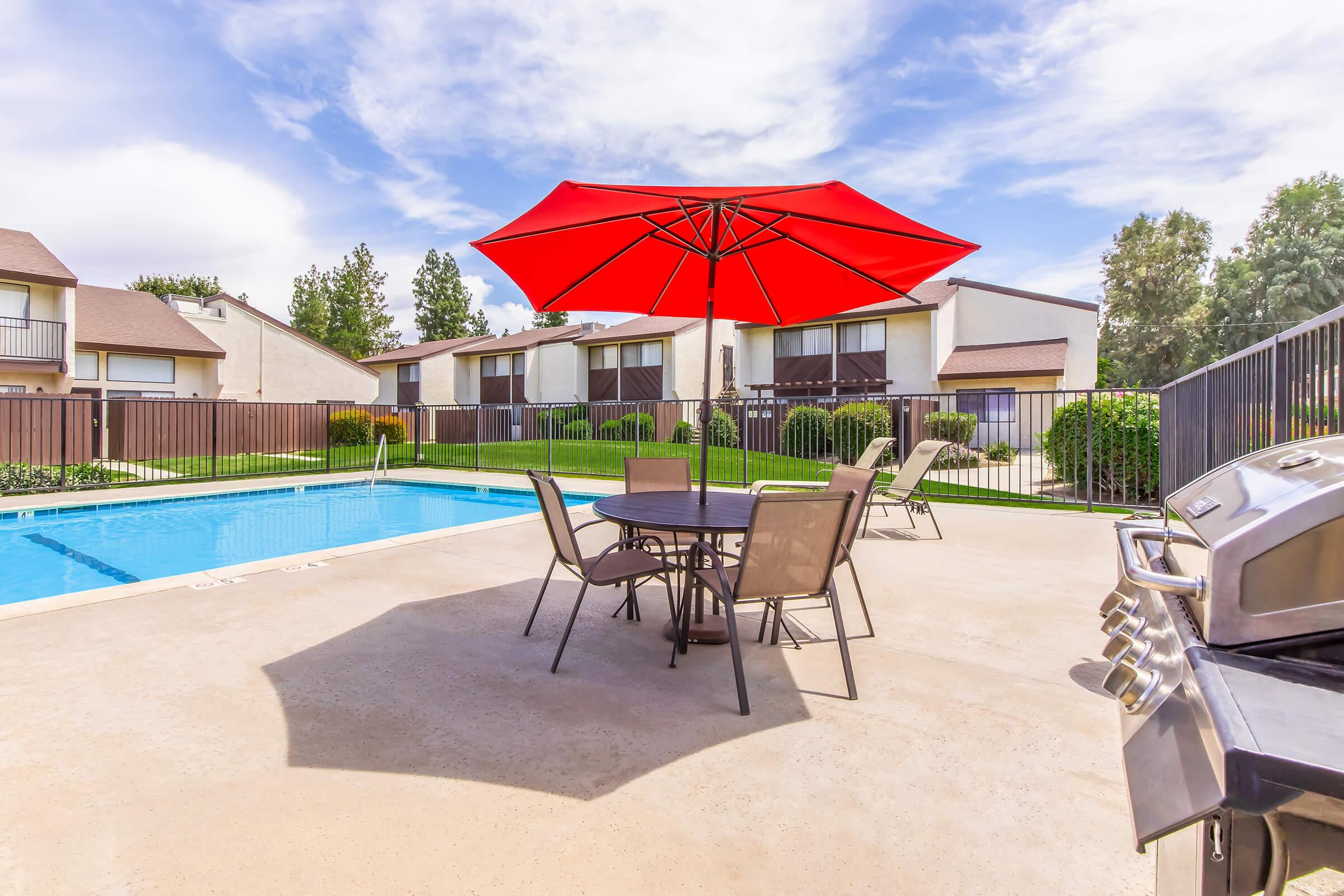A sunny outdoor pool area featuring a sparkling blue pool, surrounded by green landscaping. In the foreground, there is a patio table with four chairs beneath a large red umbrella, and a barbecue grill nearby. Apartment buildings are visible in the background under a clear blue sky with some clouds.