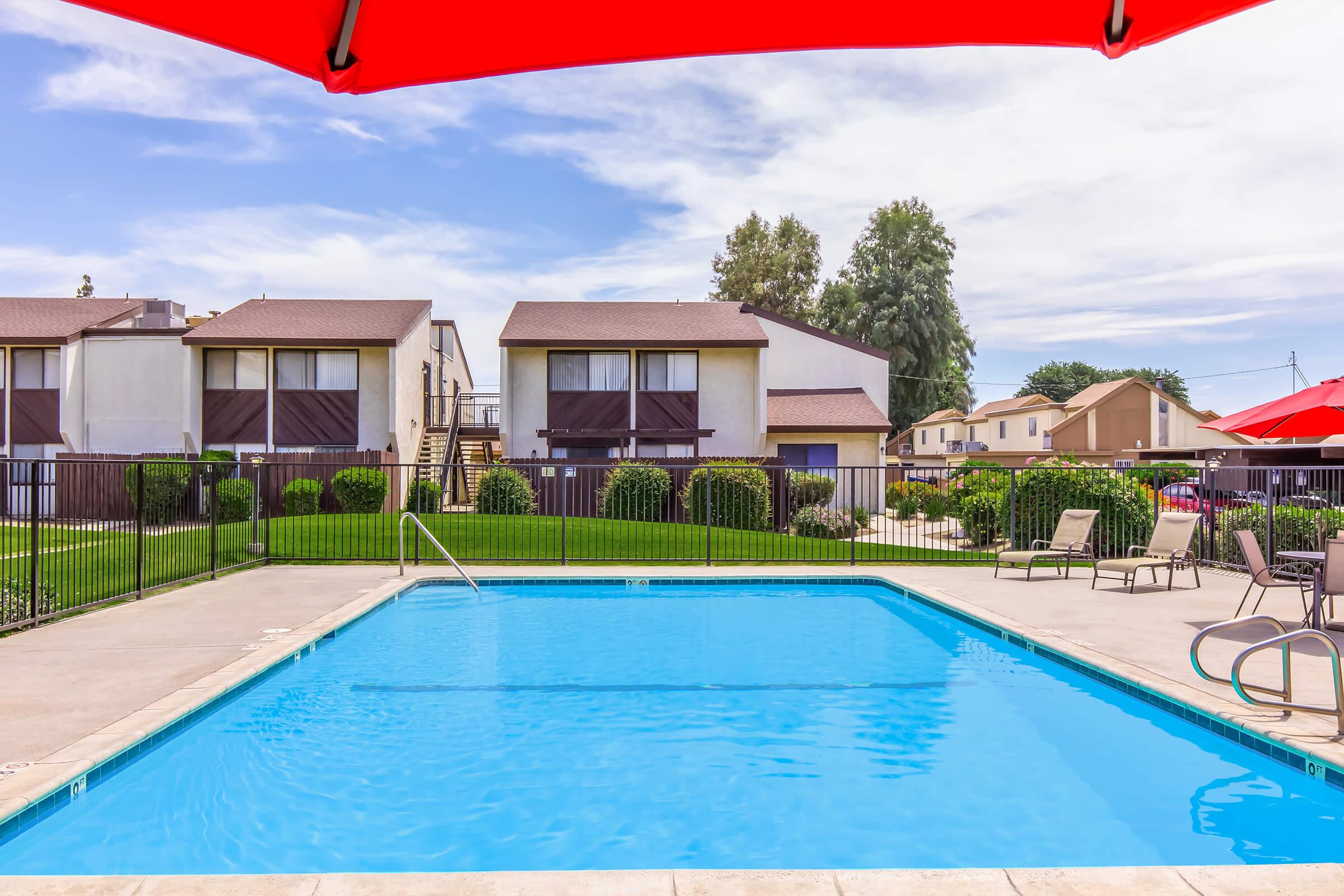 A bright outdoor swimming pool surrounded by a fenced area, featuring lounge chairs and shaded by a red umbrella. In the background, two multi-story apartment buildings are visible, along with well-maintained grass and trees, under a partly cloudy sky.