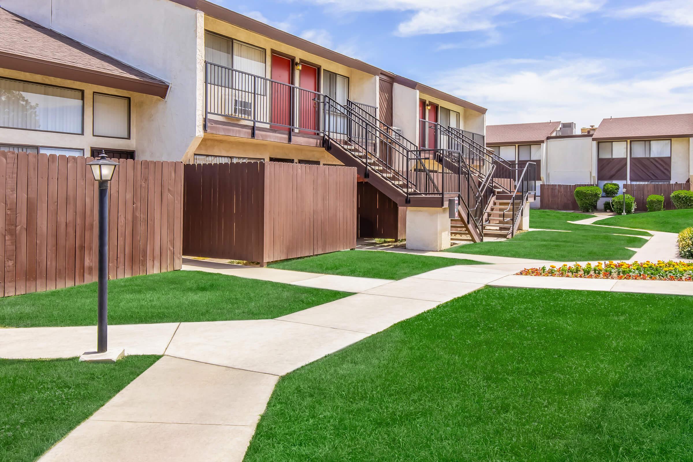 A well-maintained residential area featuring two-story apartment buildings with red doors. The grounds are landscaped with green lawns and flower beds, and a pathway leads through the area. A lamppost stands along the walkway, enhancing the inviting atmosphere.
