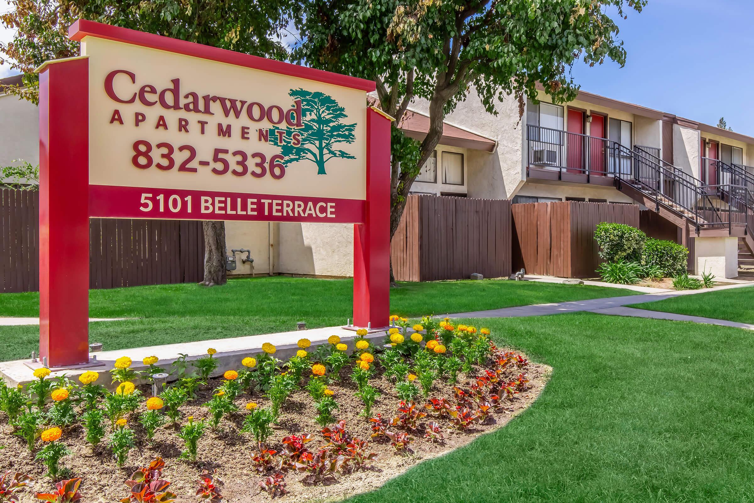 Sign for Cedarwood Apartments displaying the phone number 832-5336 and the address 5101 Belle Terrace. The sign is surrounded by colorful flowers and the apartment complex features two-storied buildings with balconies and well-maintained grass and trees in the background.