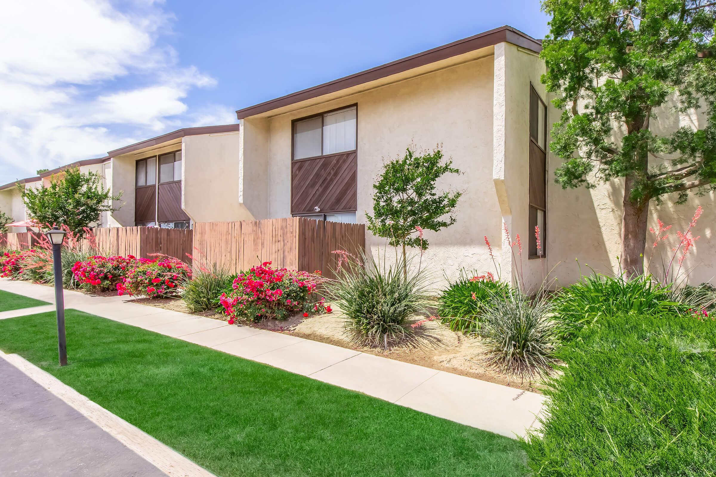 A landscaped view of a residential area featuring modern apartment buildings. The foreground includes neatly trimmed grass, flowering shrubs, and decorative plants, with a path leading to the buildings. The sky is bright with scattered clouds.