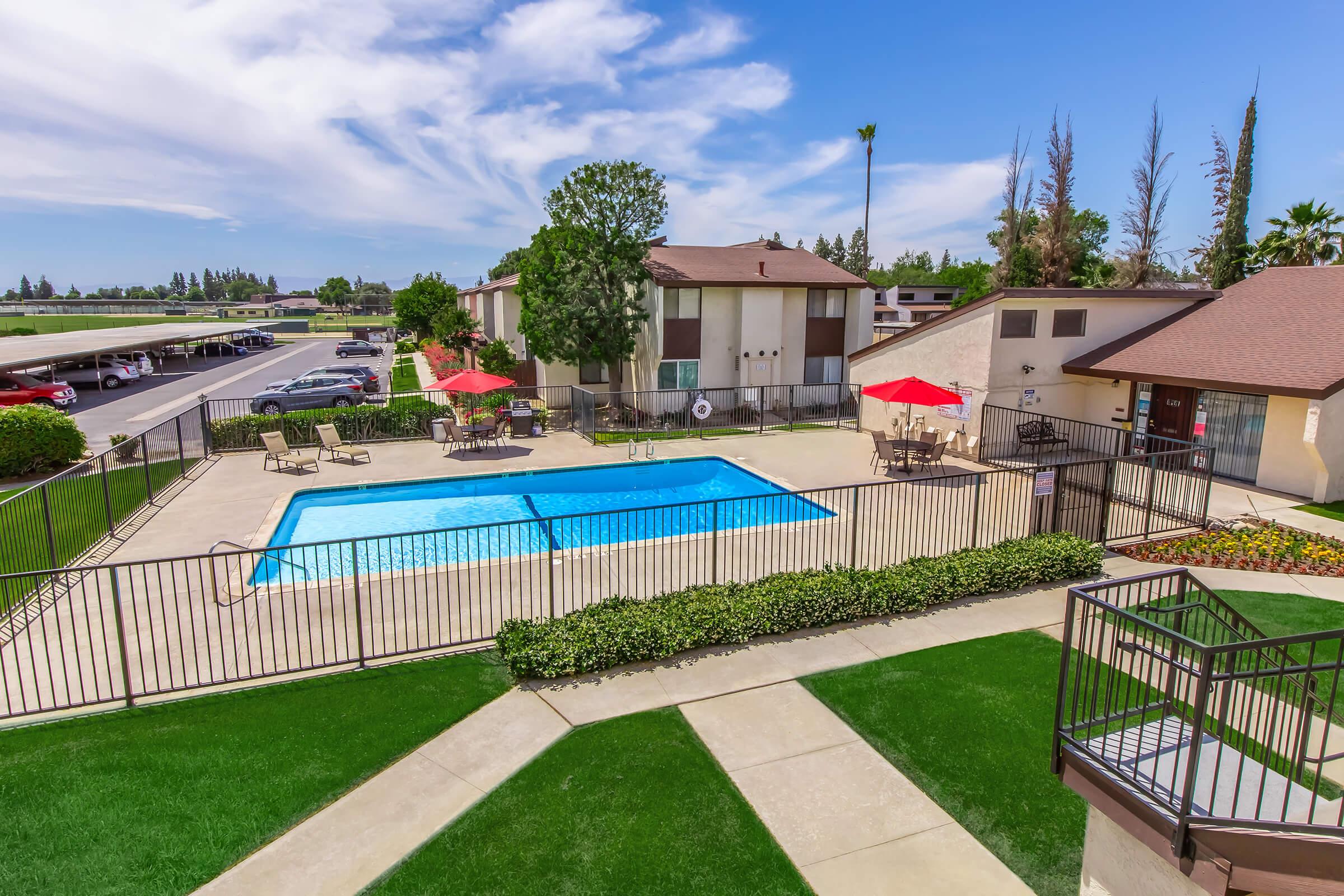 A sunlit outdoor swimming pool surrounded by a fenced area, with several lounge chairs and red umbrellas. In the background, there are residential buildings set against a clear blue sky with scattered clouds. Well-manicured lawns enhance the relaxing atmosphere.