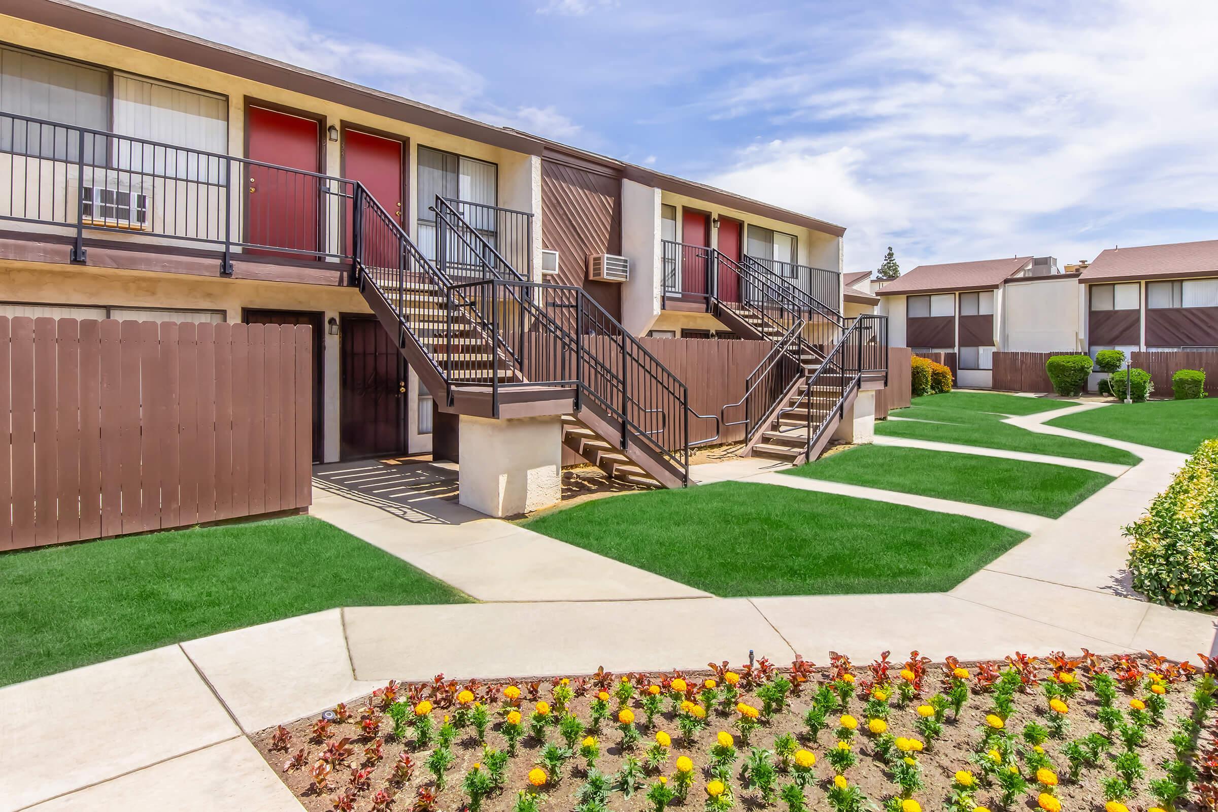 Two-story apartment complex with a landscaped green lawn and a flowerbed of yellow and red flowers in the foreground. The building features red doors with black stairs leading to the upper level. A concrete pathway winds through the well-maintained outdoor area under a partly cloudy sky.