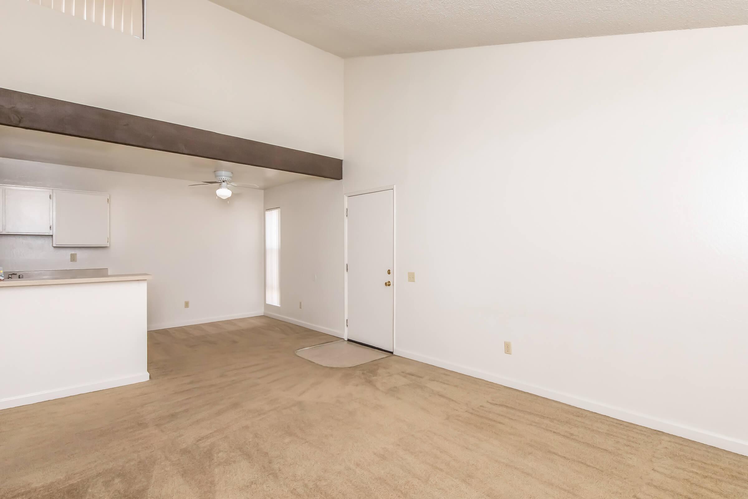 A spacious, empty room with light beige carpet and white walls, featuring a small entryway and a ceiling fan. To the left, there's a partial view of a kitchen area with light cabinetry. Bright natural light fills the space, creating an open and airy atmosphere.