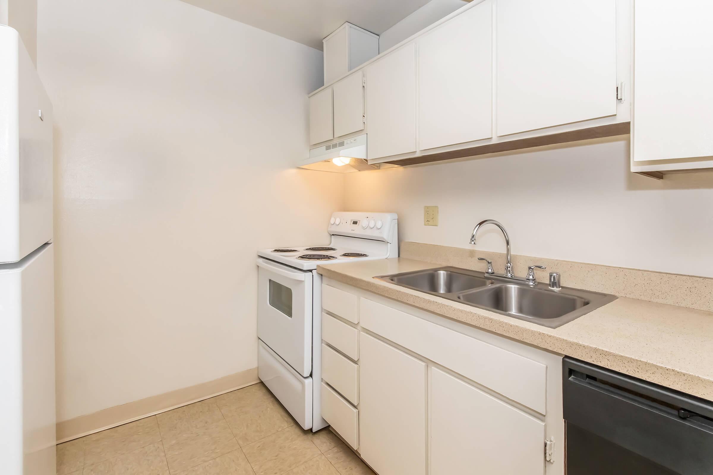 Compact kitchen featuring white cabinetry, a stove with an oven, a stainless steel sink with a faucet, and a dishwasher. The walls are painted light, and the floor is a light beige color. A refrigerator is also visible on the left side of the image.