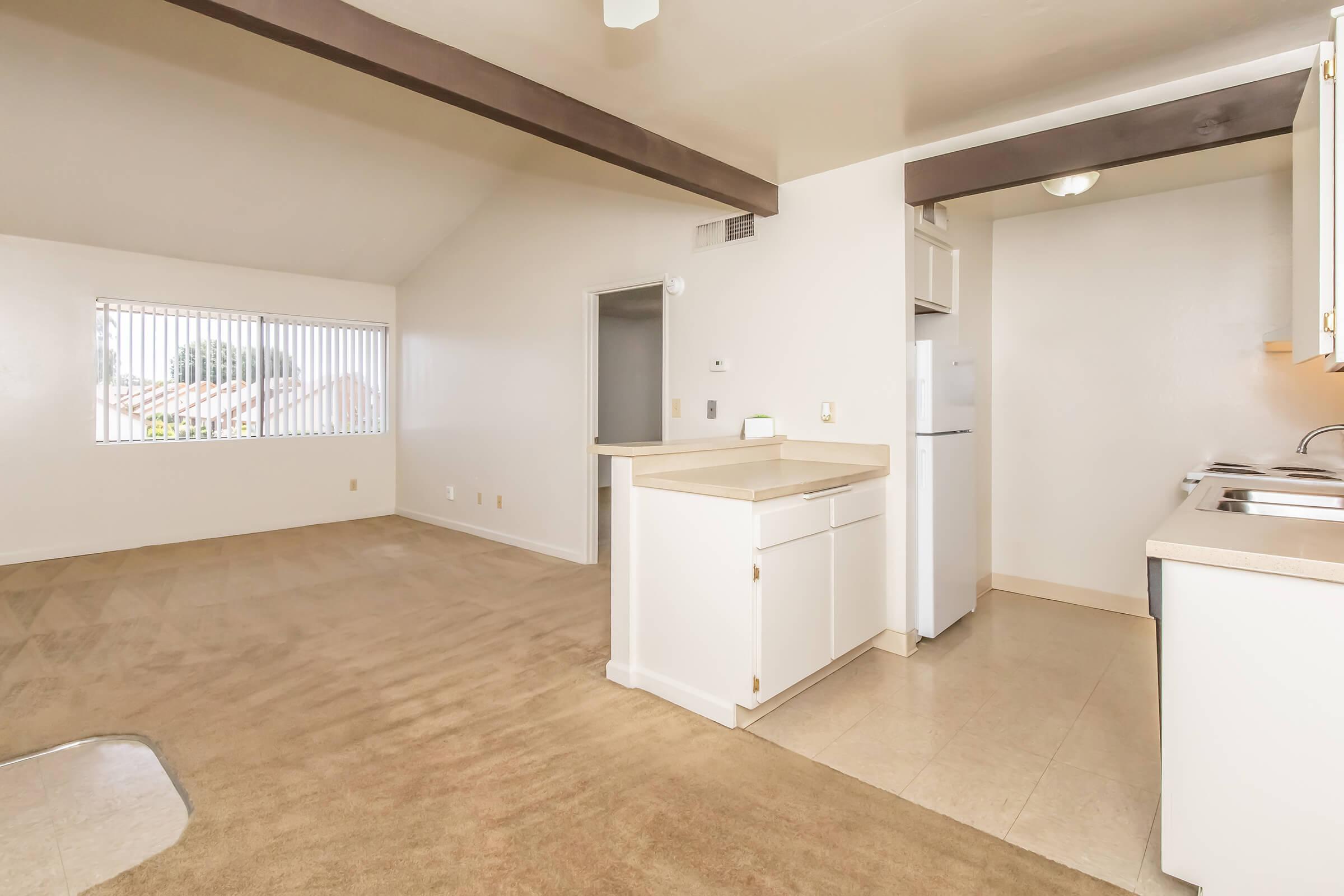 A spacious, empty room with beige carpet and white walls. To the right, there is a small kitchen area with a countertop, sink, and appliances. A window with vertical blinds allows natural light, revealing a view outside. The ceiling features wooden beams, adding character to the space.