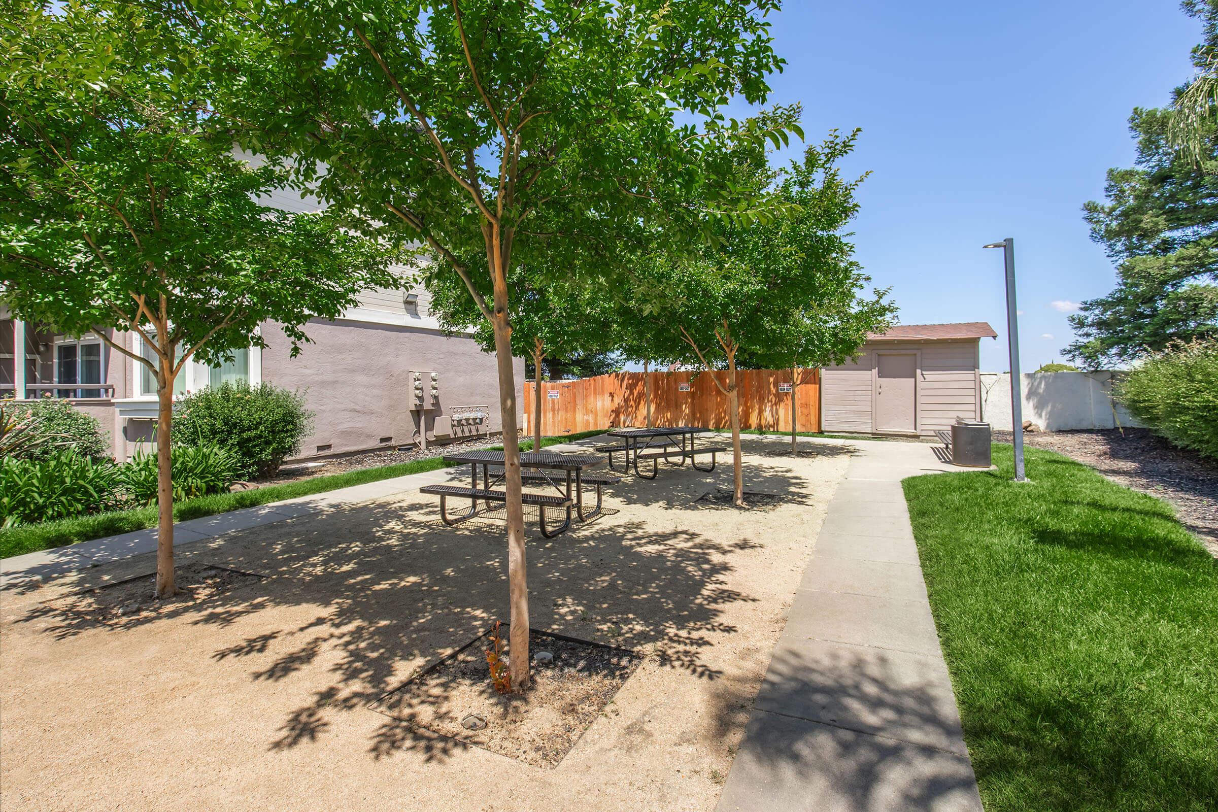 A landscaped outdoor area featuring several picnic tables under green trees, with a gravel surface. A brown wooden fence and a small storage shed are visible in the background, along with patches of grass and shrubs around the space. The sky is clear and blue, indicating a sunny day.