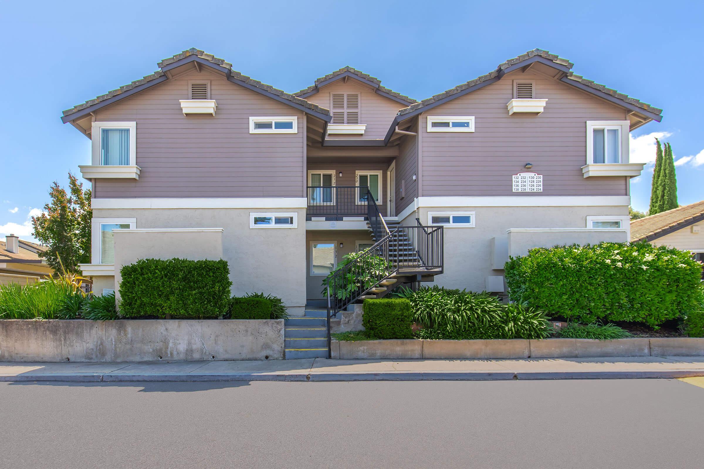 A multi-story residential building with a light purple exterior and a dark roof. It features multiple balconies and a staircase leading to the entrance. The landscape includes well-maintained shrubs and small trees, set against a clear blue sky.