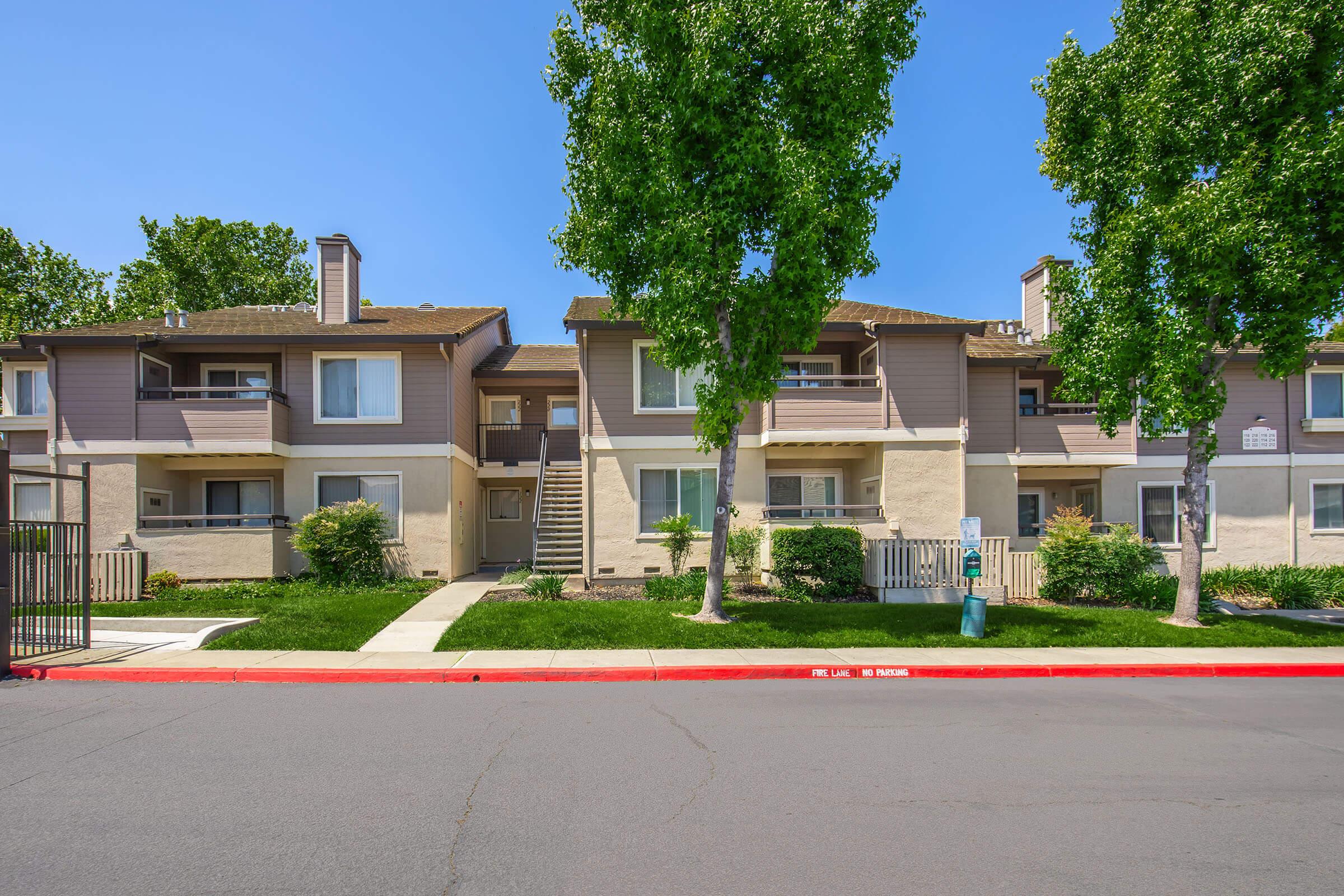 A sunny exterior view of a residential apartment complex, featuring two-story buildings with balconies, surrounded by green grass and trees. A paved walkway leads to the entrance, with a red curb and a mailbox visible in the foreground. Clear blue sky in the background.