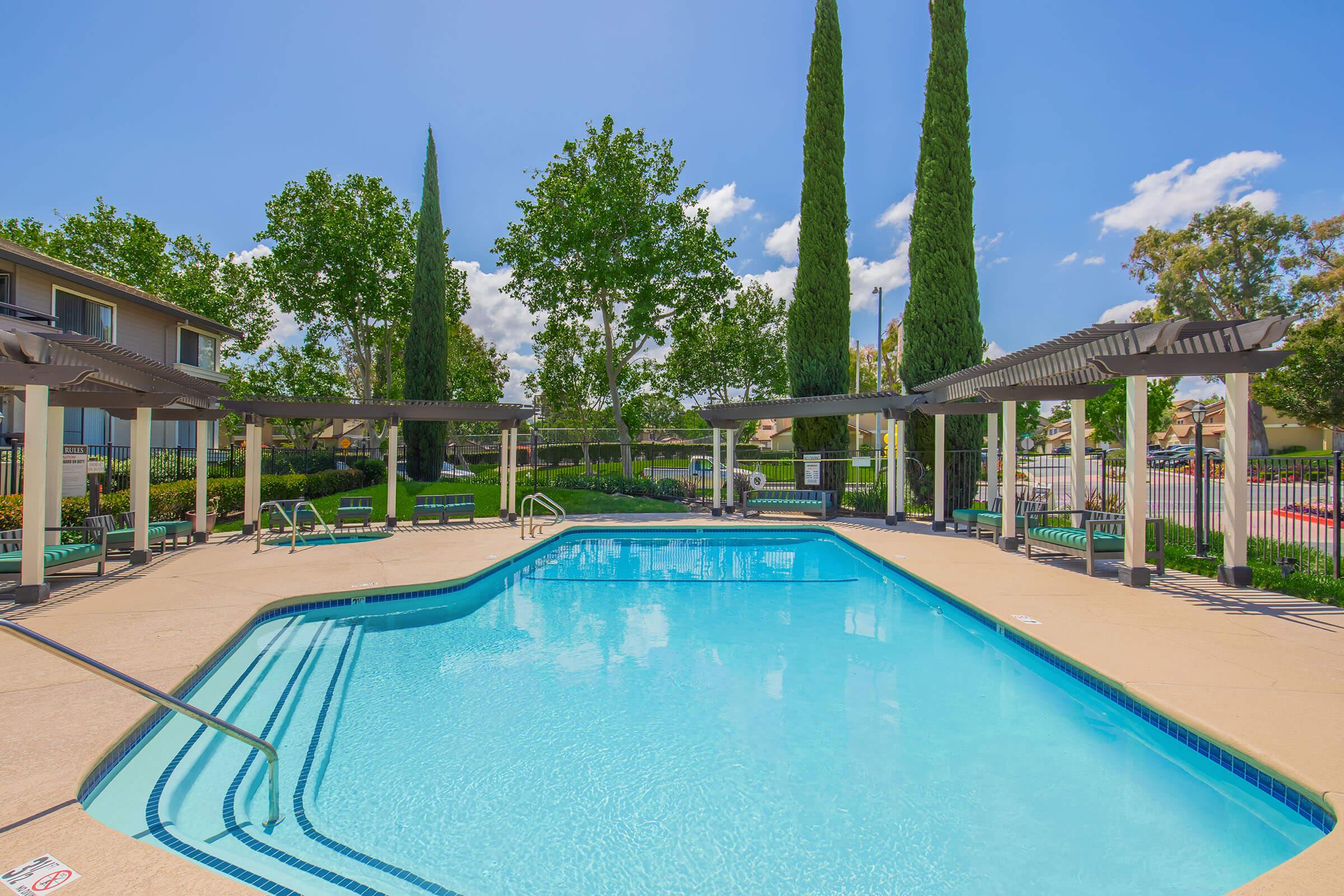 A bright, clear swimming pool surrounded by lounge chairs and shaded pergolas. Towering green trees in the background and a blue sky with fluffy clouds enhance the serene atmosphere. The pool area is well-maintained, offering a relaxing outdoor space for leisure and recreation.