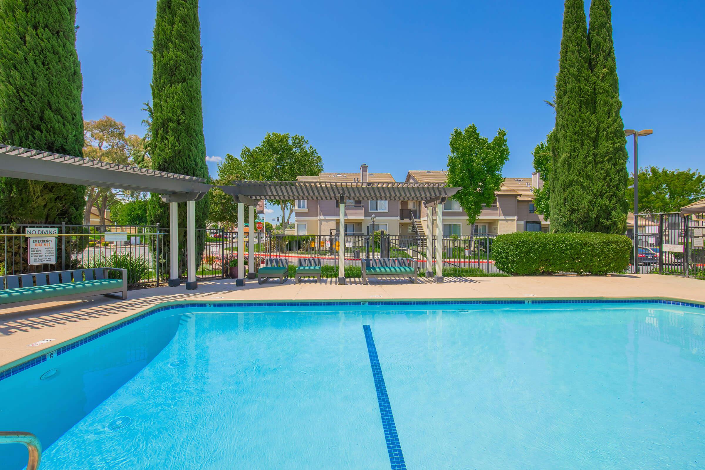 A clear blue swimming pool surrounded by green trees and landscaped areas. There are pergolas and lounge seating nearby, with a fence enclosing the pool area. The background features residential buildings, creating a serene community atmosphere under a sunny sky.