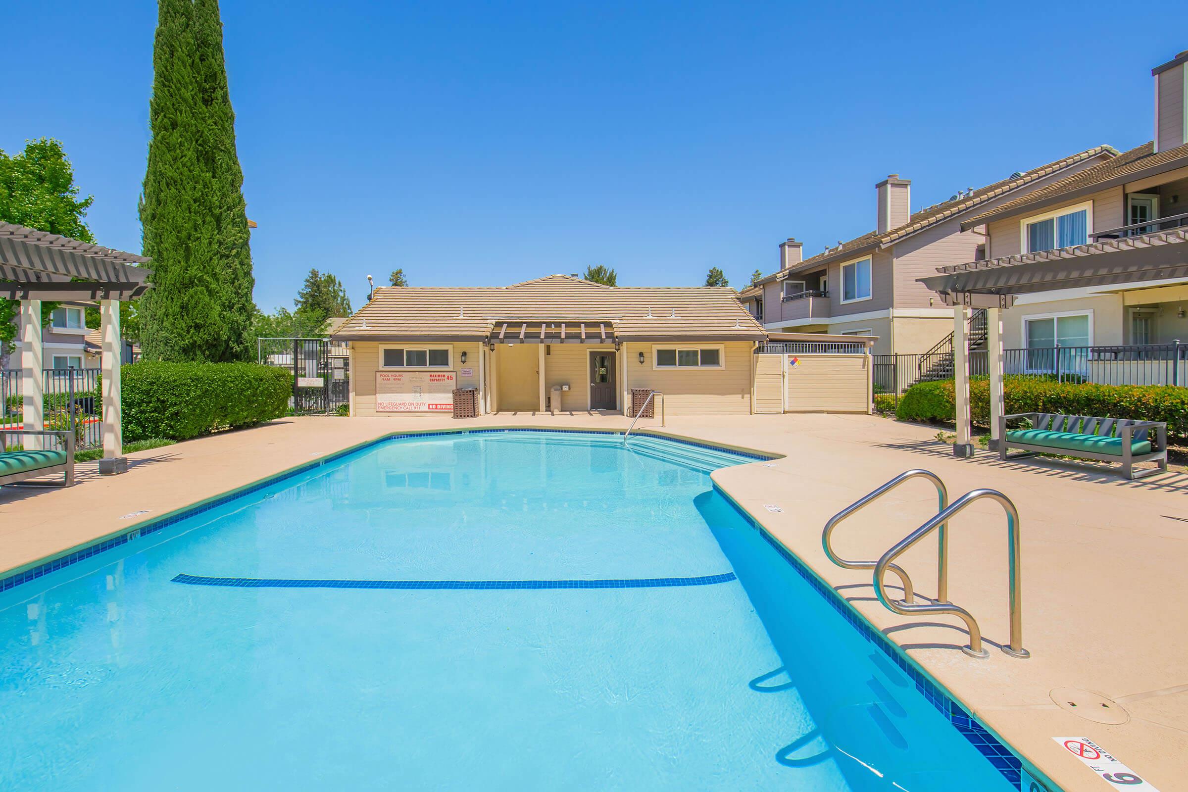 A clear blue swimming pool surrounded by a sunlit deck. Nearby, a small building with a roof provides amenities. Lush greenery and trees are in the background, with several residential buildings visible, creating a welcoming and relaxing atmosphere.